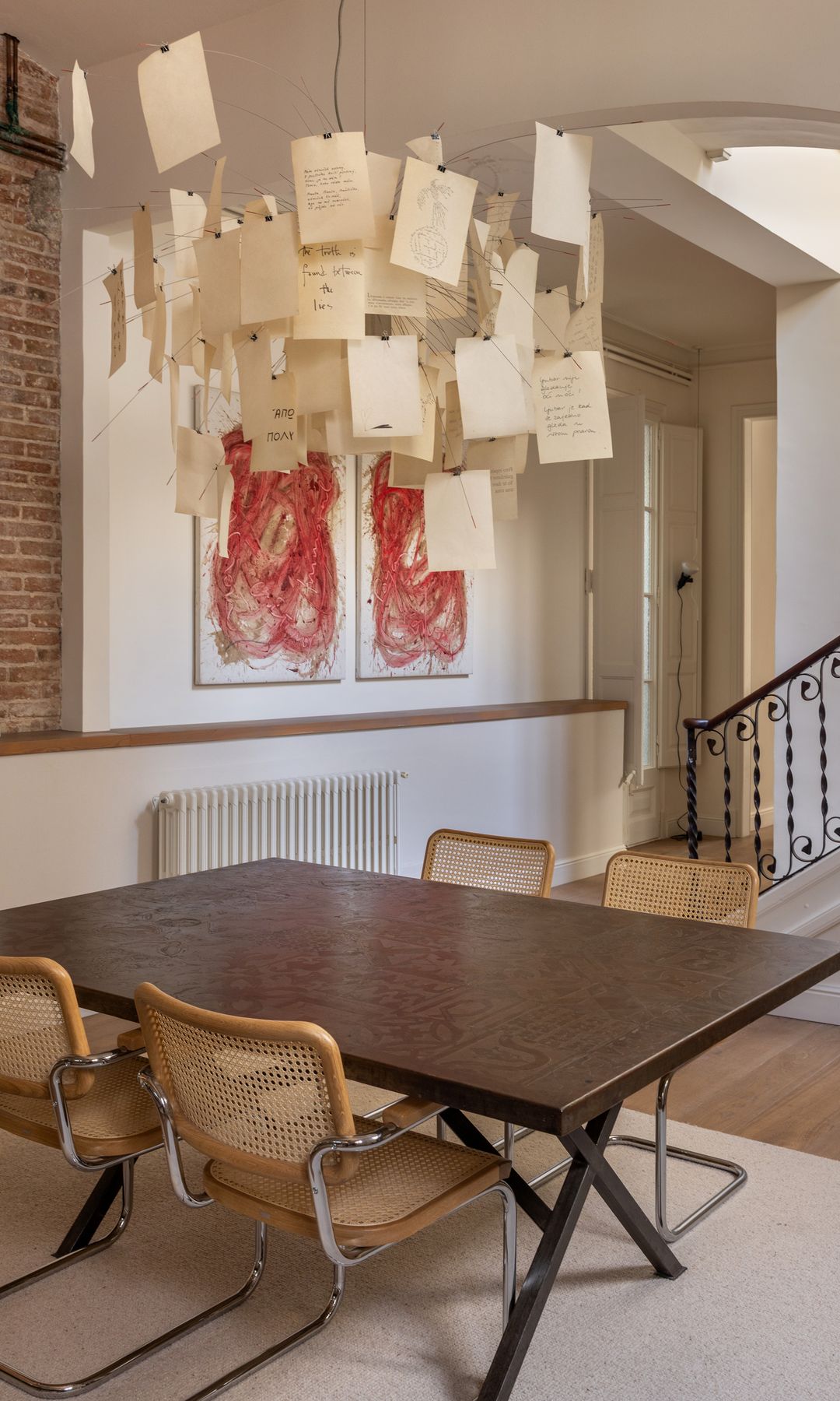Comedor con lámpara de techo con mensajes de papel, mesa de madera, sillas Cesca, alfombra blanca, pared de ladrillo, cuadros en la pared