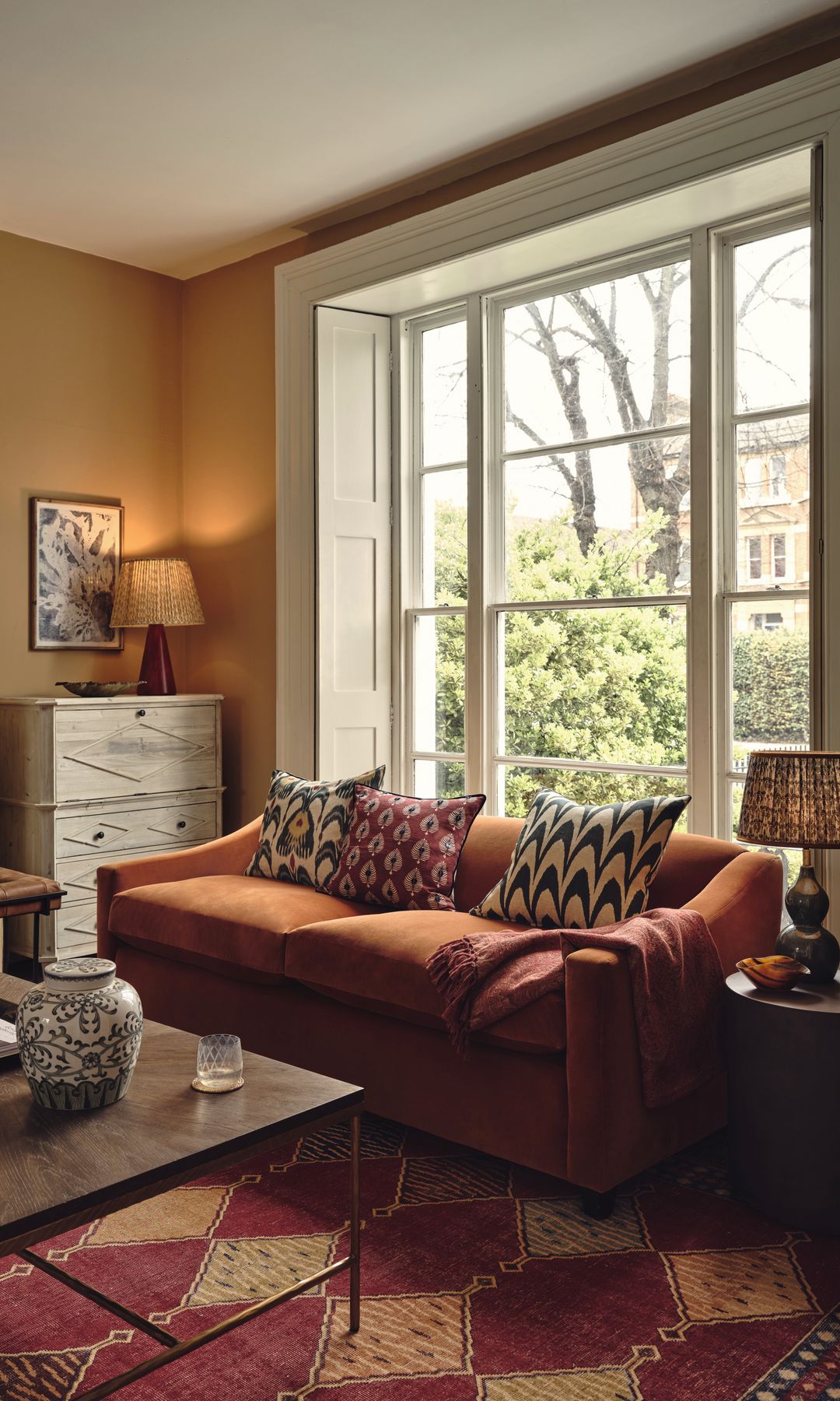 Salón con chimenea, gran ventanal, sofá marrón bajo la ventana, alfombra con motivos geométricos burdeos, aparador alto, lámpara de sobremesa, mesa de centro