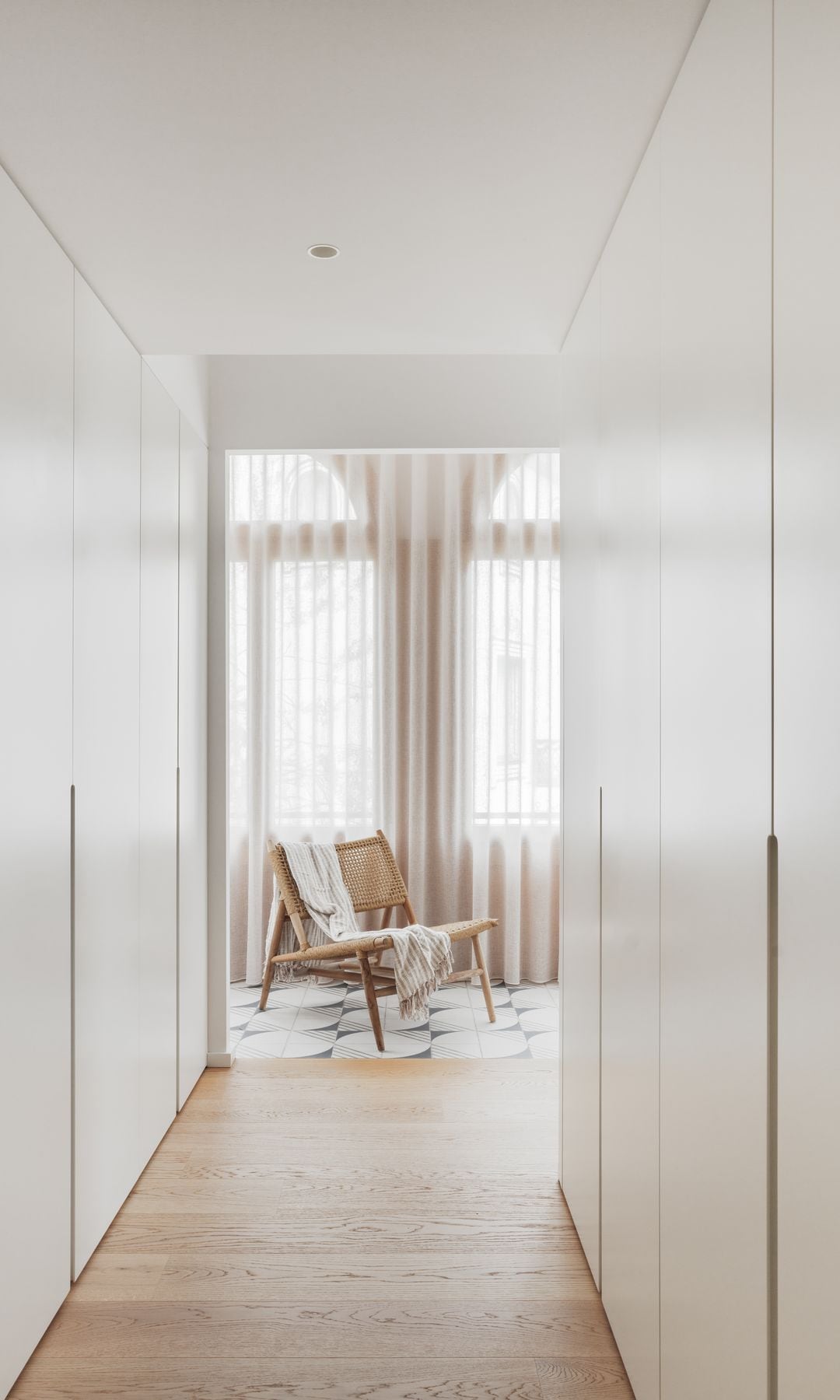 Pasillo vestidor con armarios blancos de suelo a techo, suelo de madera y sillón de fibras vegetales