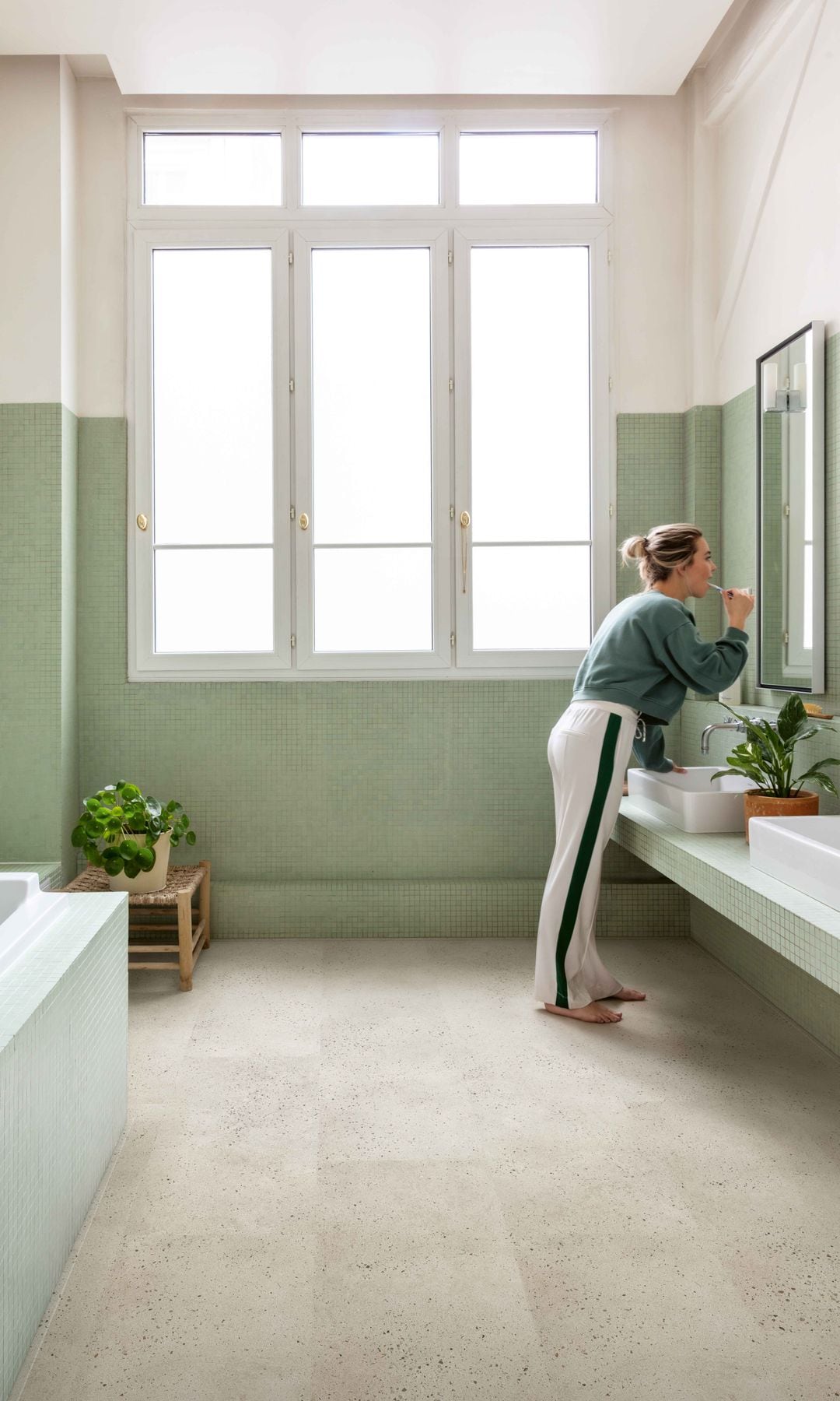 Mujer descalza lavándose los dientes en baño con paredes verdes, bañera y suelo laminado con efecto hormigón 
