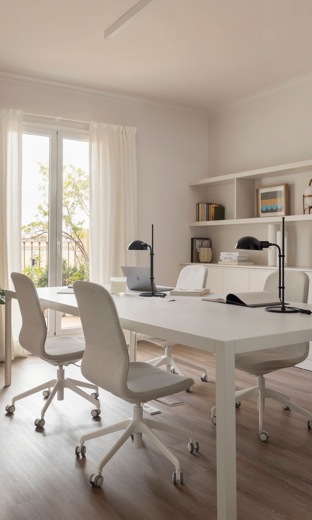 Despacho en casa con mesa de trabajo XL de color blanco, cuatro sillas de oficina blancas con ruedas, dos flexos negros, estantería blanca con lámpara Cesta y libros, cortinas blancas