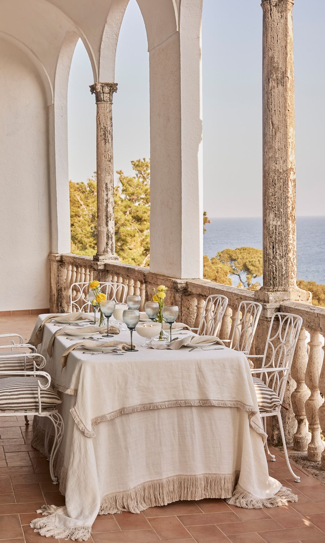 Mesa con mantel beige en terraza con vistas al mar, sillas de hierro blanco