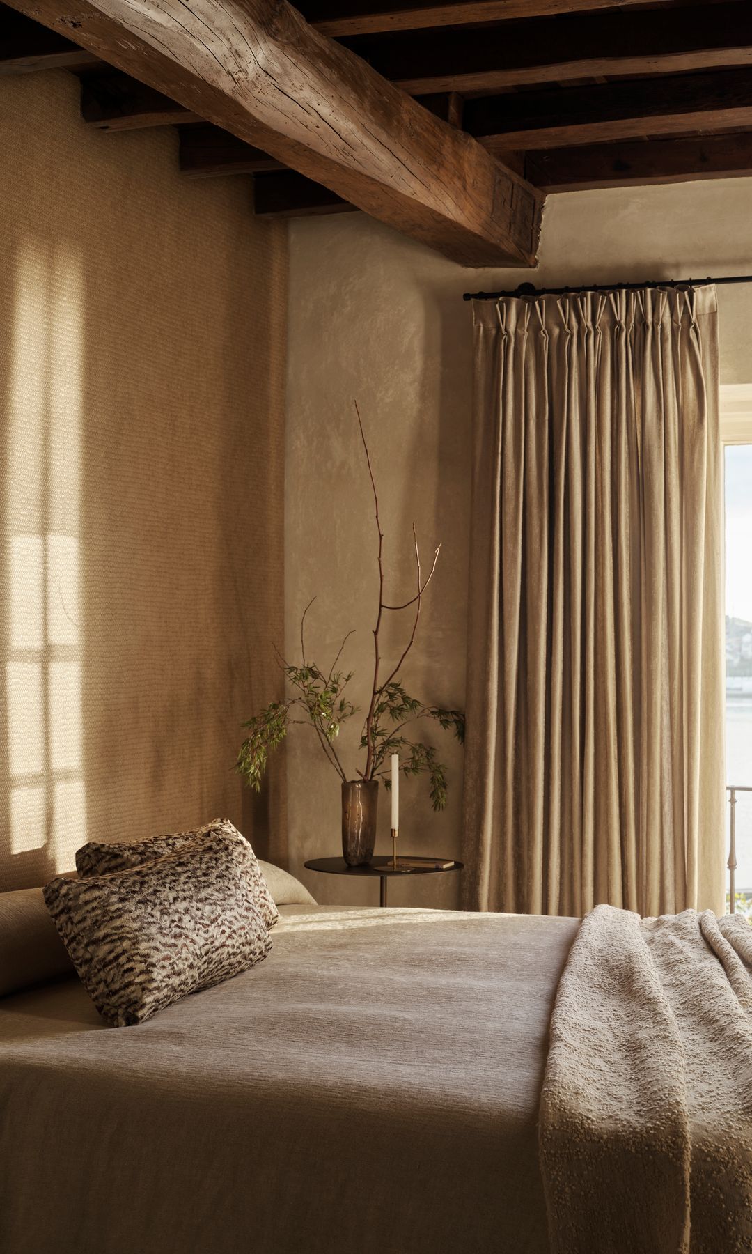 Dormitorio con cortinas beiges, ropa de cama en tonos tierra, vigas de madera, ventanal con salida al balcón 