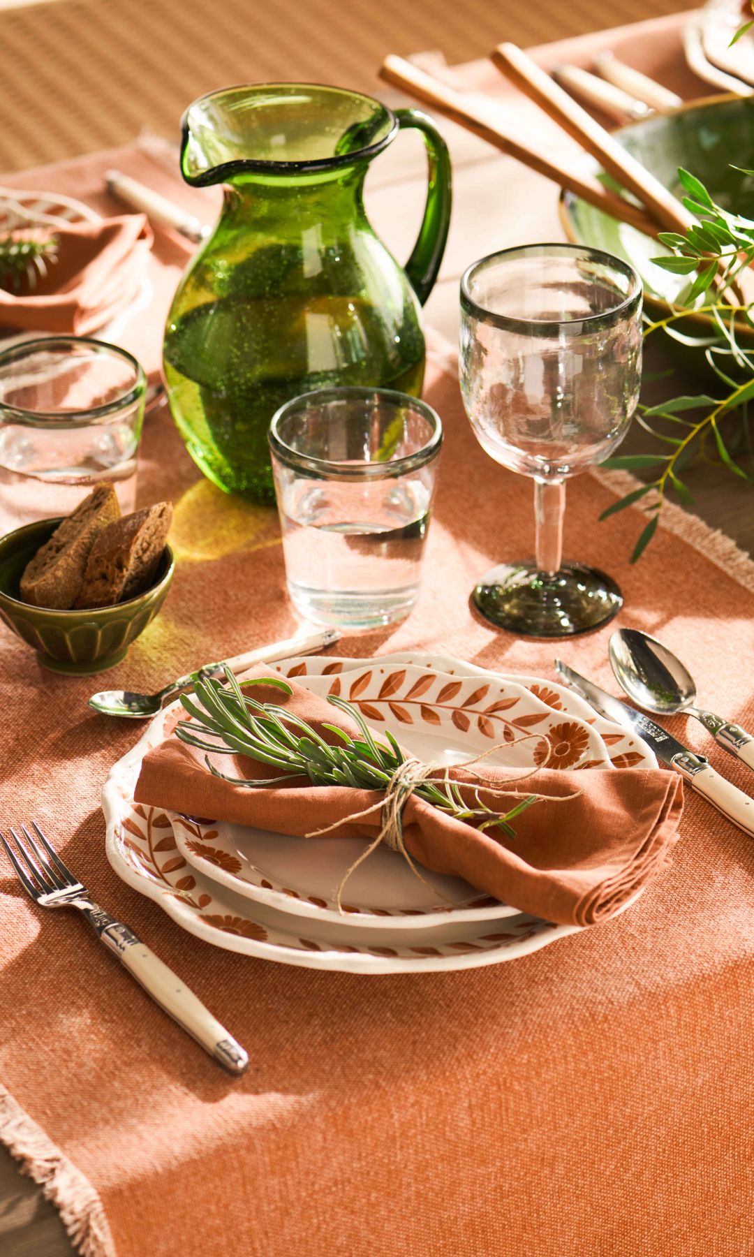 Mesa de verano con manteles individuales, vajilla blanca con motivos naranjas, jarra de cristal verde, cubiertos de hueso