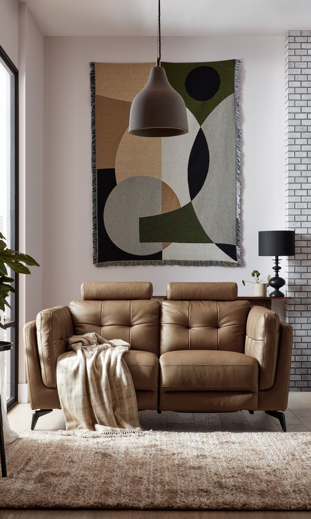Sofá de cuero marrón con asientos reclinables, manta sobre el sofá, lámpara de techo, alfombra beige, tapiz en la pared, lámpara de sobremesa negra 