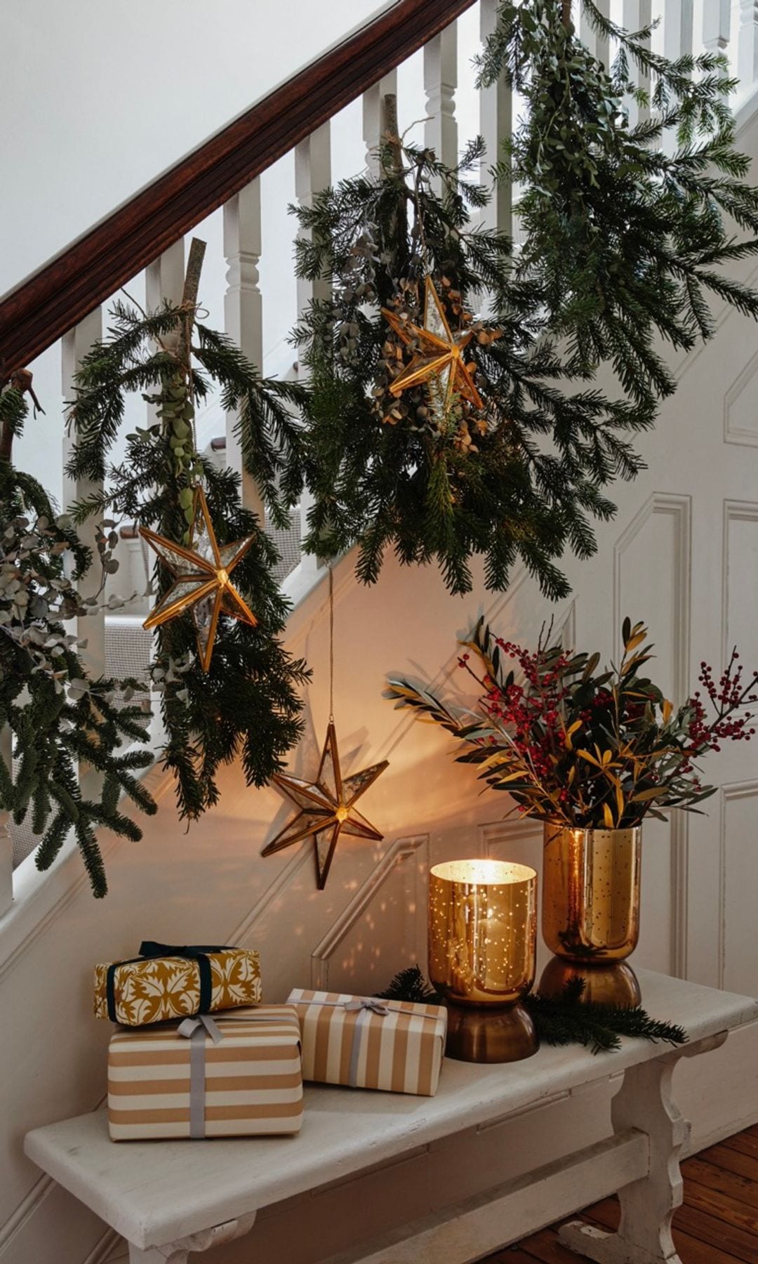 Recibidor con ramas de abeto colgadas en la barandilla, banco blanco con regalos, velas y arreglo floral en soporte dorado