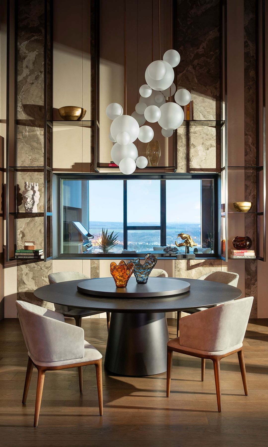 Comedor con mesa redonda negra, lámpara de techo con globos blancos, medio tabique de cristal comunica con cocina exterior