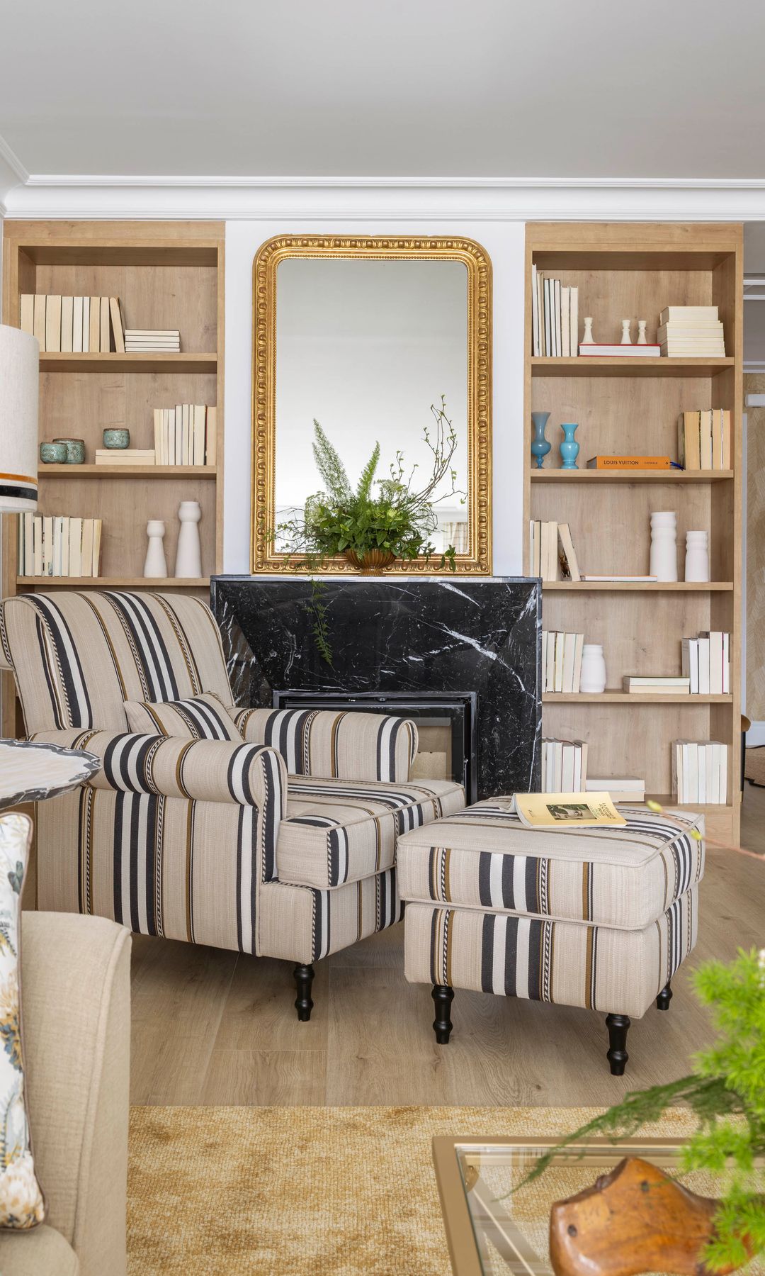 Salón con chimenea de mármol negro, espejo con el marco dorado, librerías de madera, sillón de rayas con reposapiés a juego, libro abierto 