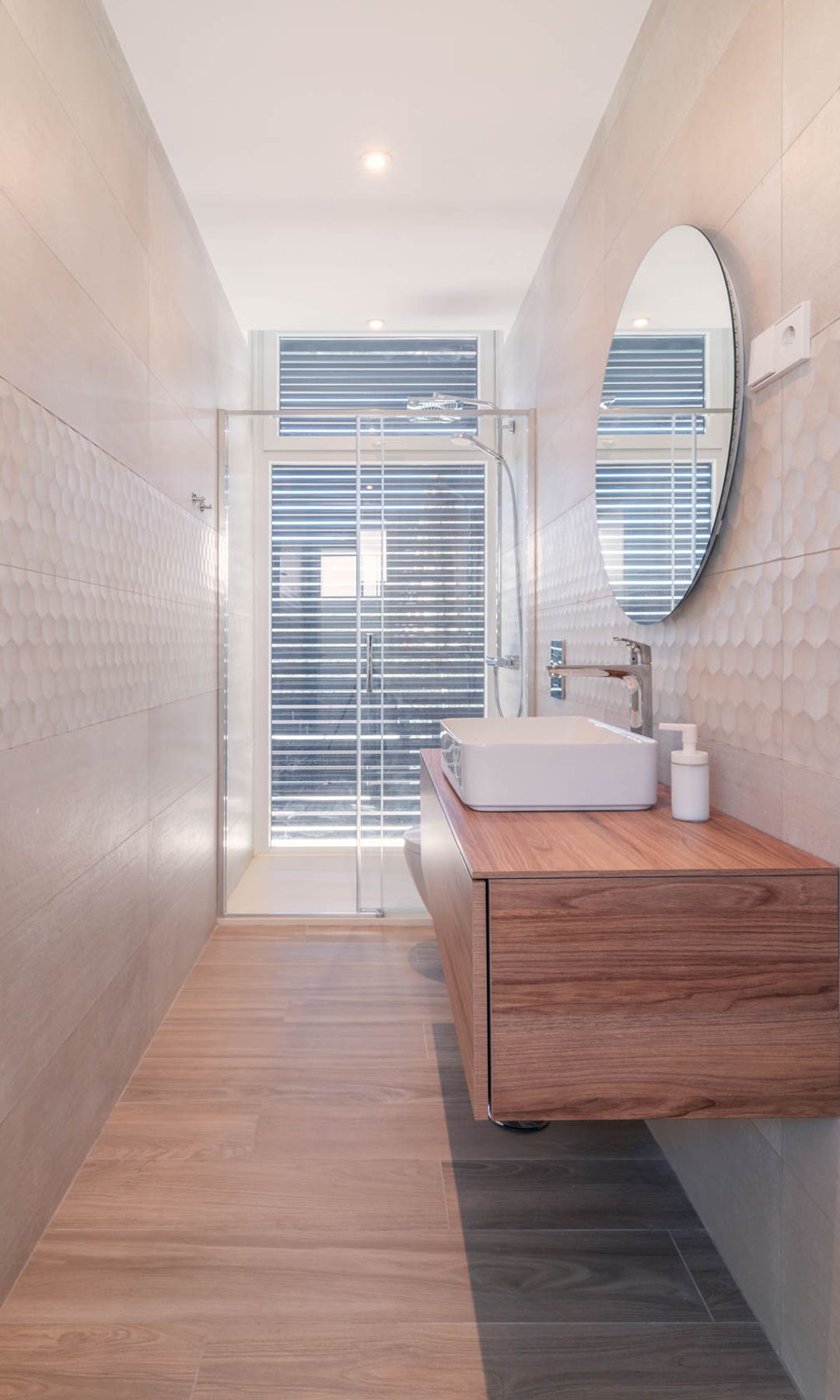 Baño con ducha, de planta estrecha, mueble de lavabo suspendido de madera, espejo redondo, lavabo sobre encimer, mampara de ducha