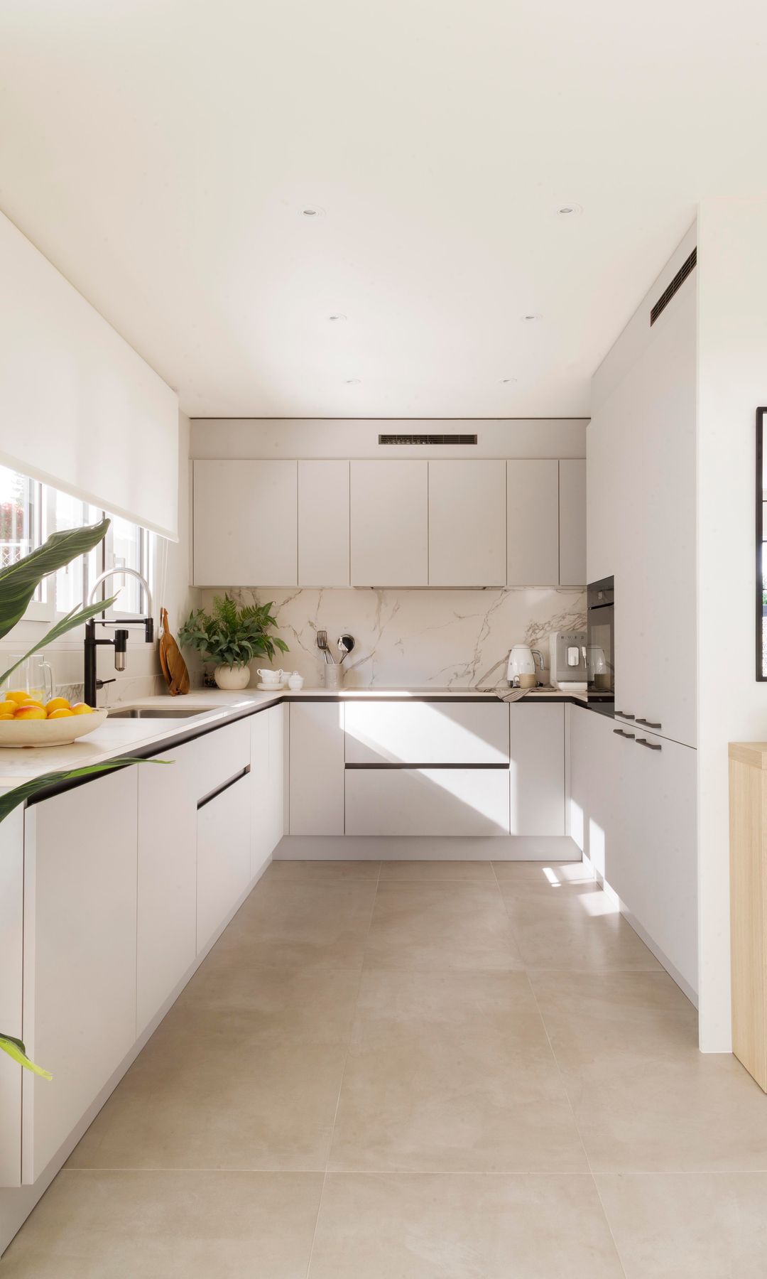 Cocina blanca en u, con tiradores negros, suelo beige, estor blanco, plantas en la encimera