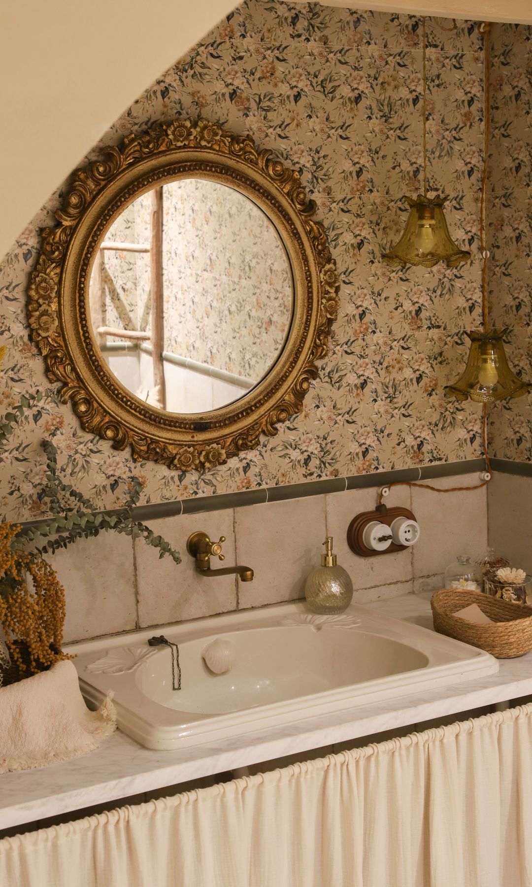 Baño con papel pintado de flores, espejo dorado, cortinilla en lugar de cajones en el mueble de lavabo, apliques de pared, grifería dorada
