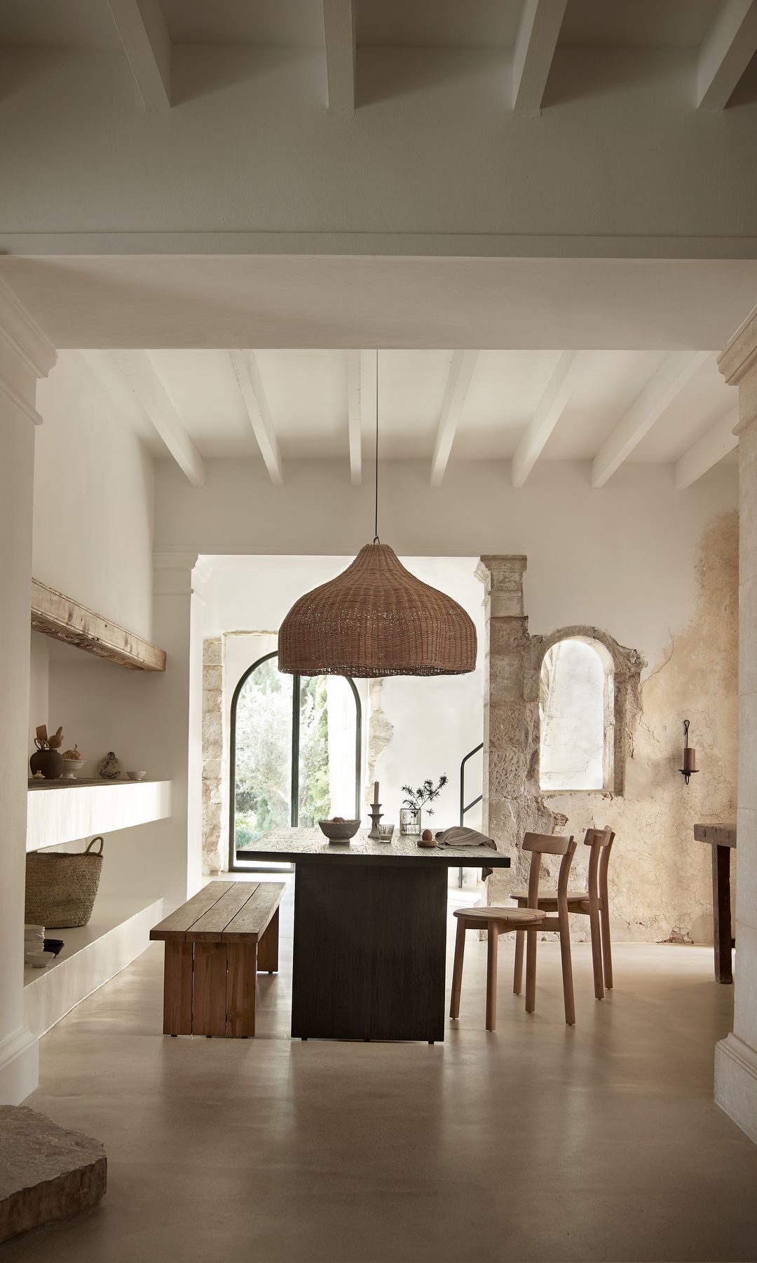 Comedor con banco de madera, mesa negra, lámpara de techo de fibras vegetales, sillas, vigas en el techo, estantería de obra 