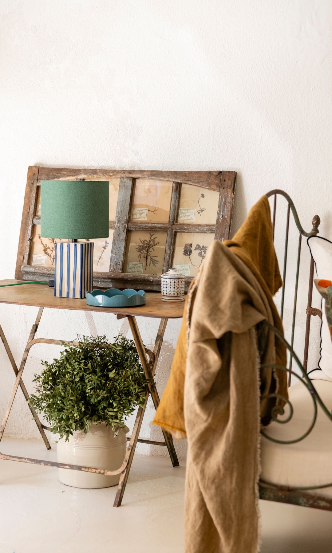Mesa de tijera con maceta con planta debajo, lámpara de sobremesa encima, cuadro de láminas con marco de madera, vacíabolsillos azul