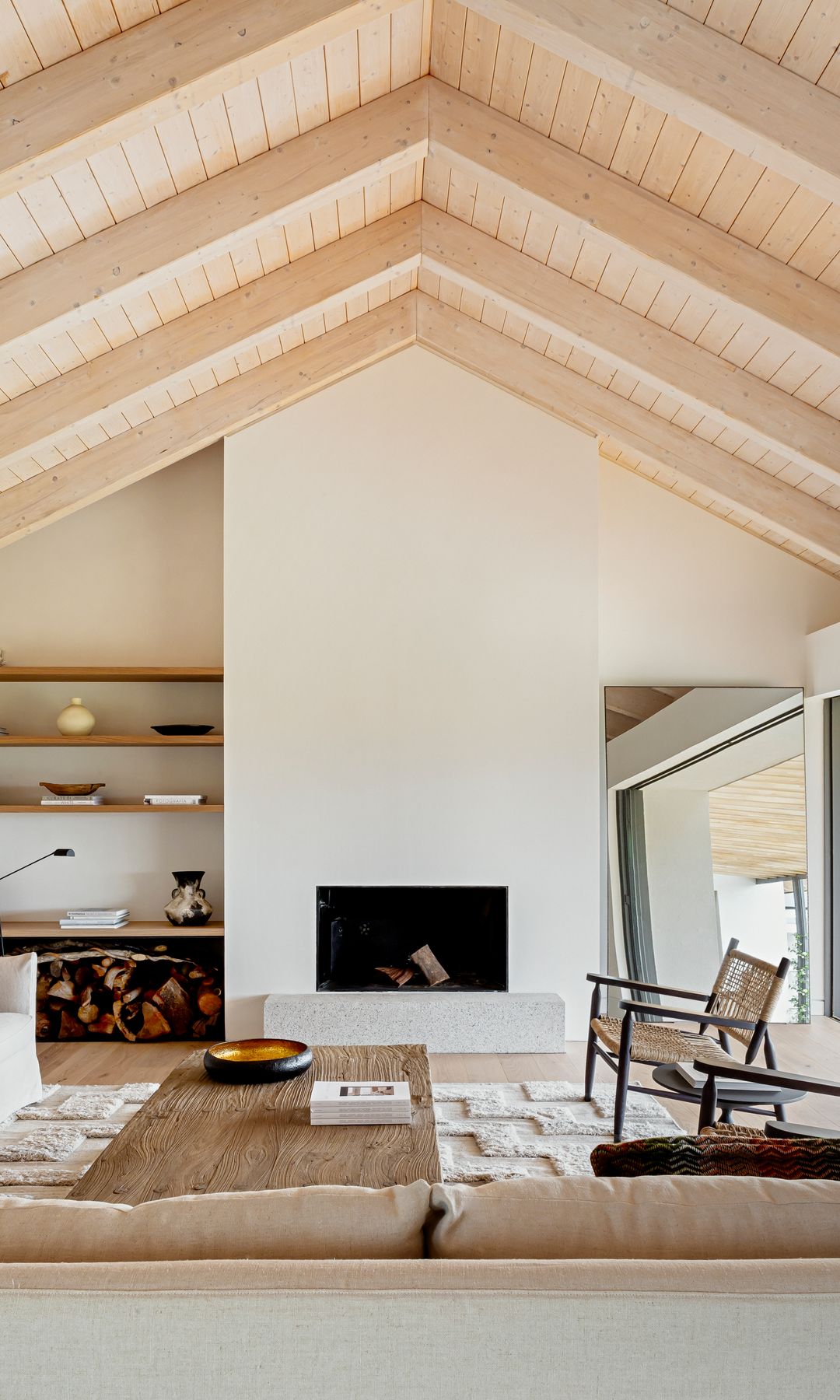Salón con chimenea y techo de madera, estantería de obra con baldas de madera, leñera, sofás blancos, alfombra, mesa de centro de madera, sillas de madera