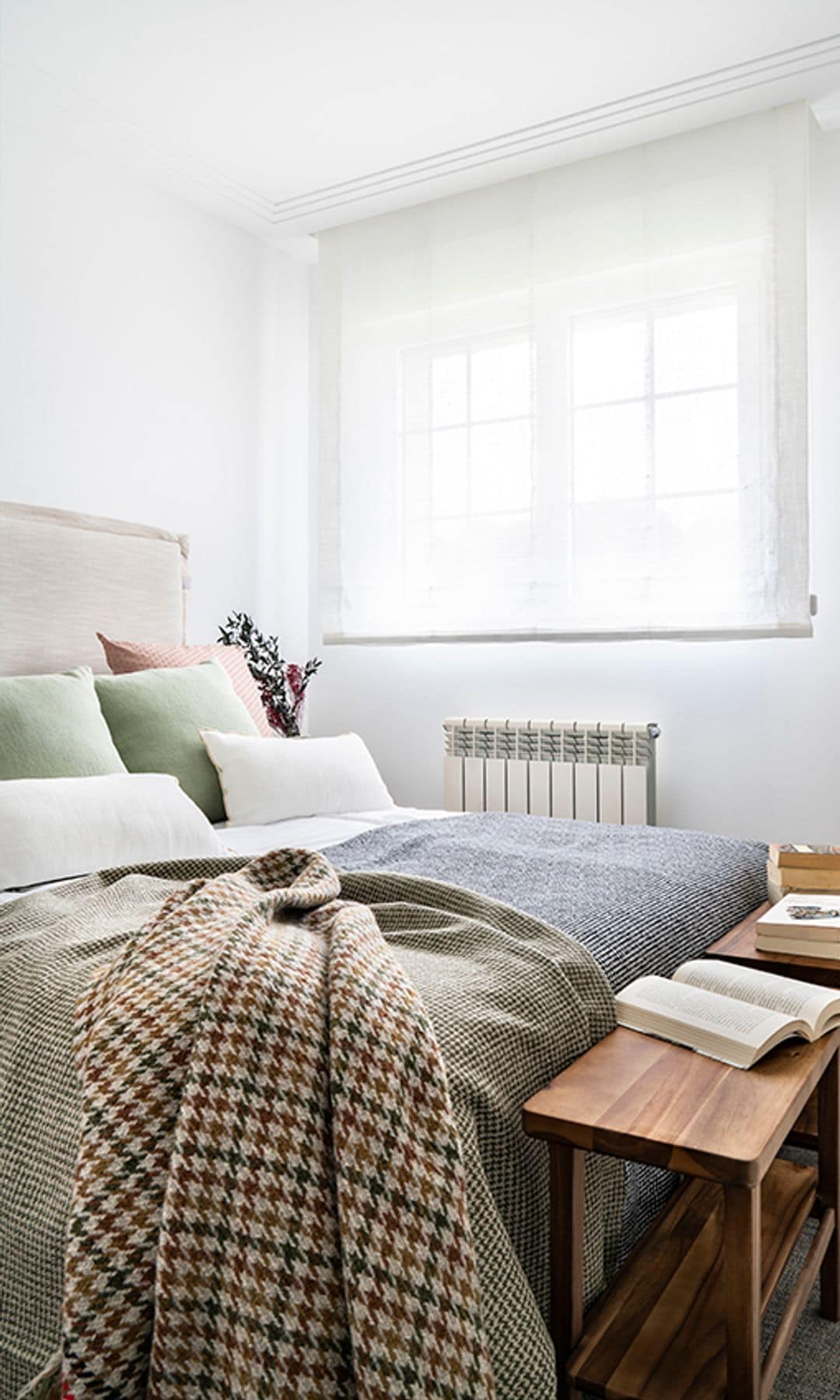 Dormitorio con estor blanco, cabecero tapizado, ropa de cama de cuadros y pata de gallo, banqueta de madera a los pies de la cama, espejo de pie con el marco de madera