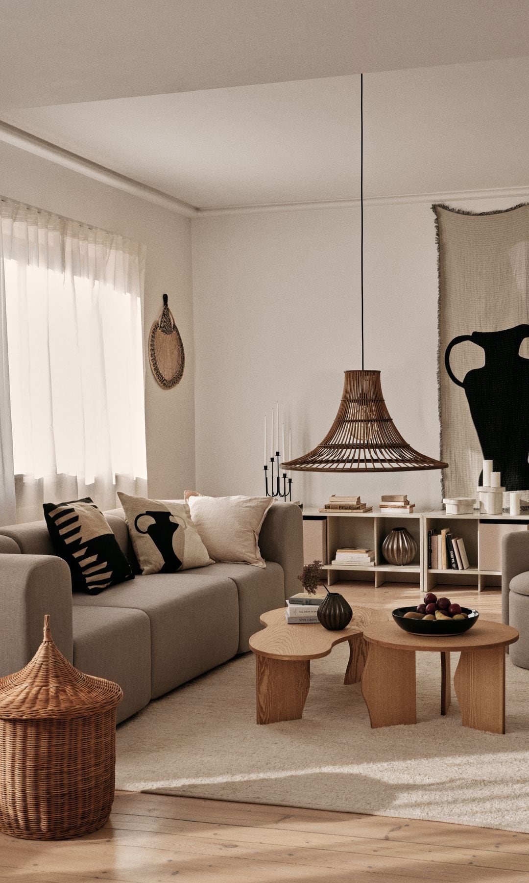 Salón cozy en tonos tierra con sofá beige, butaca beige, alfombra de pelo, mesas de centro de madera, cesto de fibras vegetales, tapiz en la pared, cortinas blancas, lámpara de techo de fibras vegetales