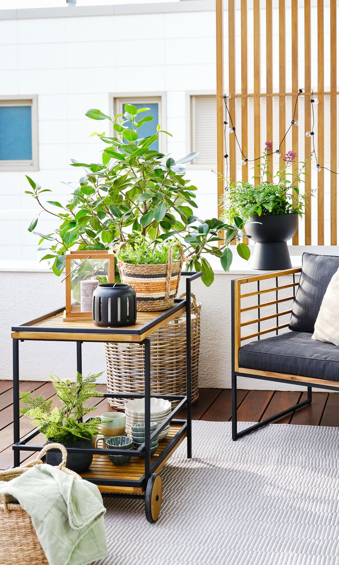 Terraza con muebles de madera y aluminio negro, carrito auxiliar con ruedas, alfombra y plantas