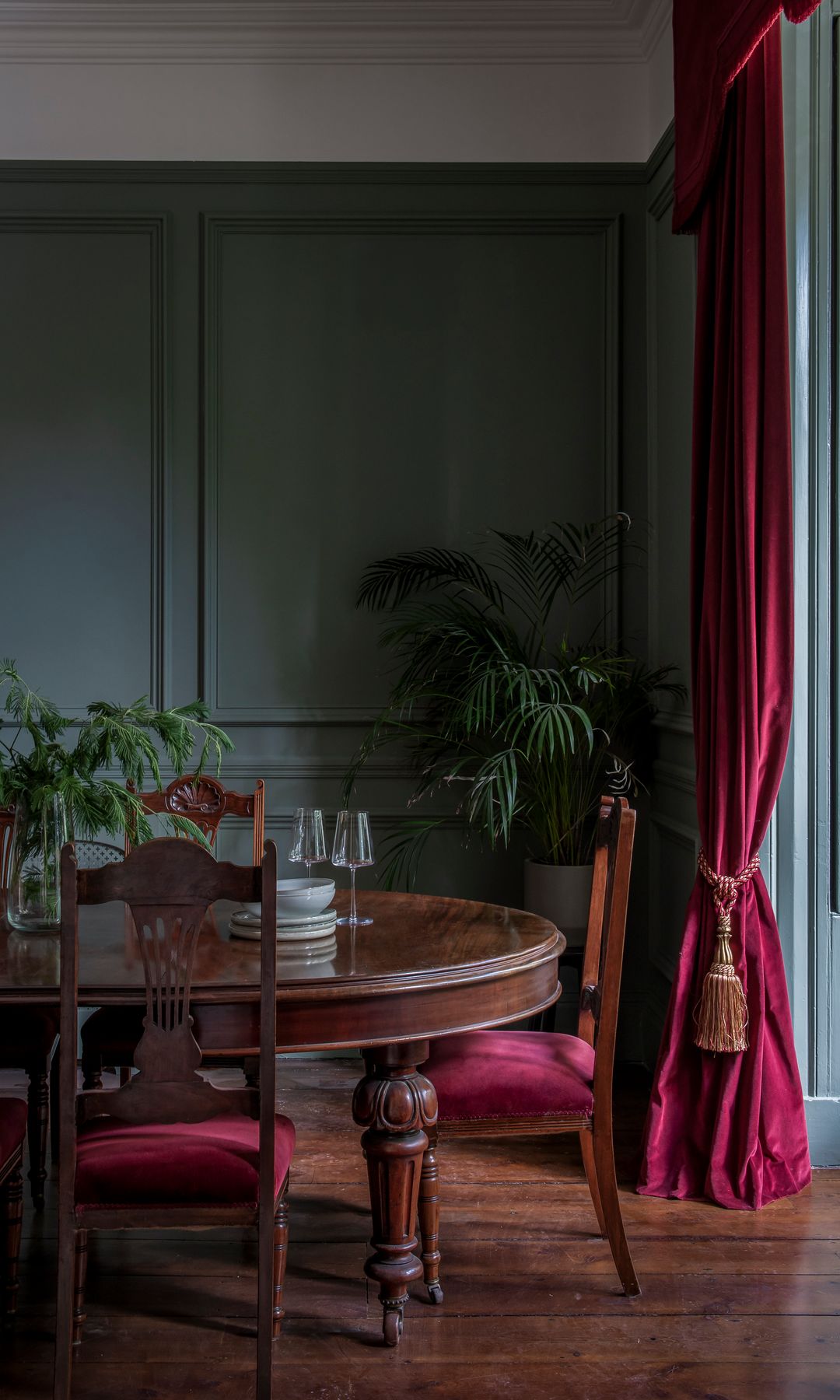 Comedor con mesa redonda de madera antigua, molduras grises en las paredes, cortinas de terciopelo en color rojos, sillas tapizadas en rojo oscuro 