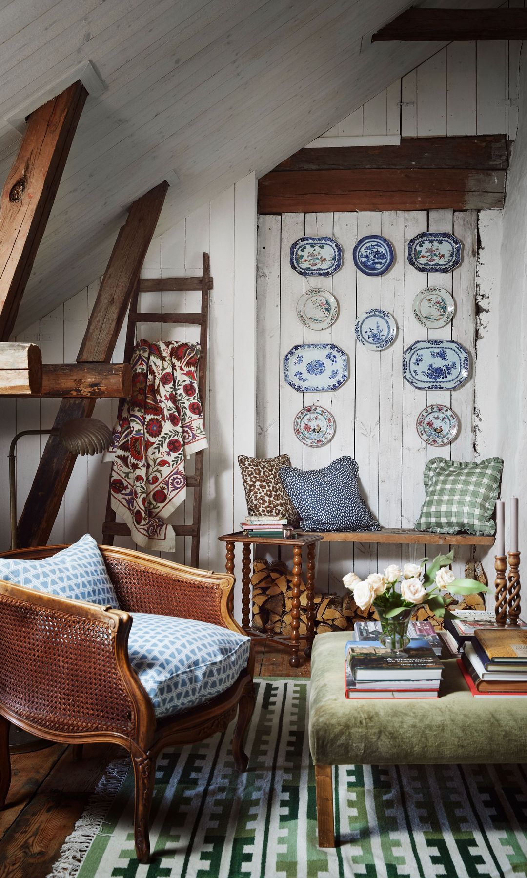 Salón con butaca con asiento de cuadros azules y blancos, platos en la pared, mesa de centro tapizada, banco, cojines, libros, jarrón con flores