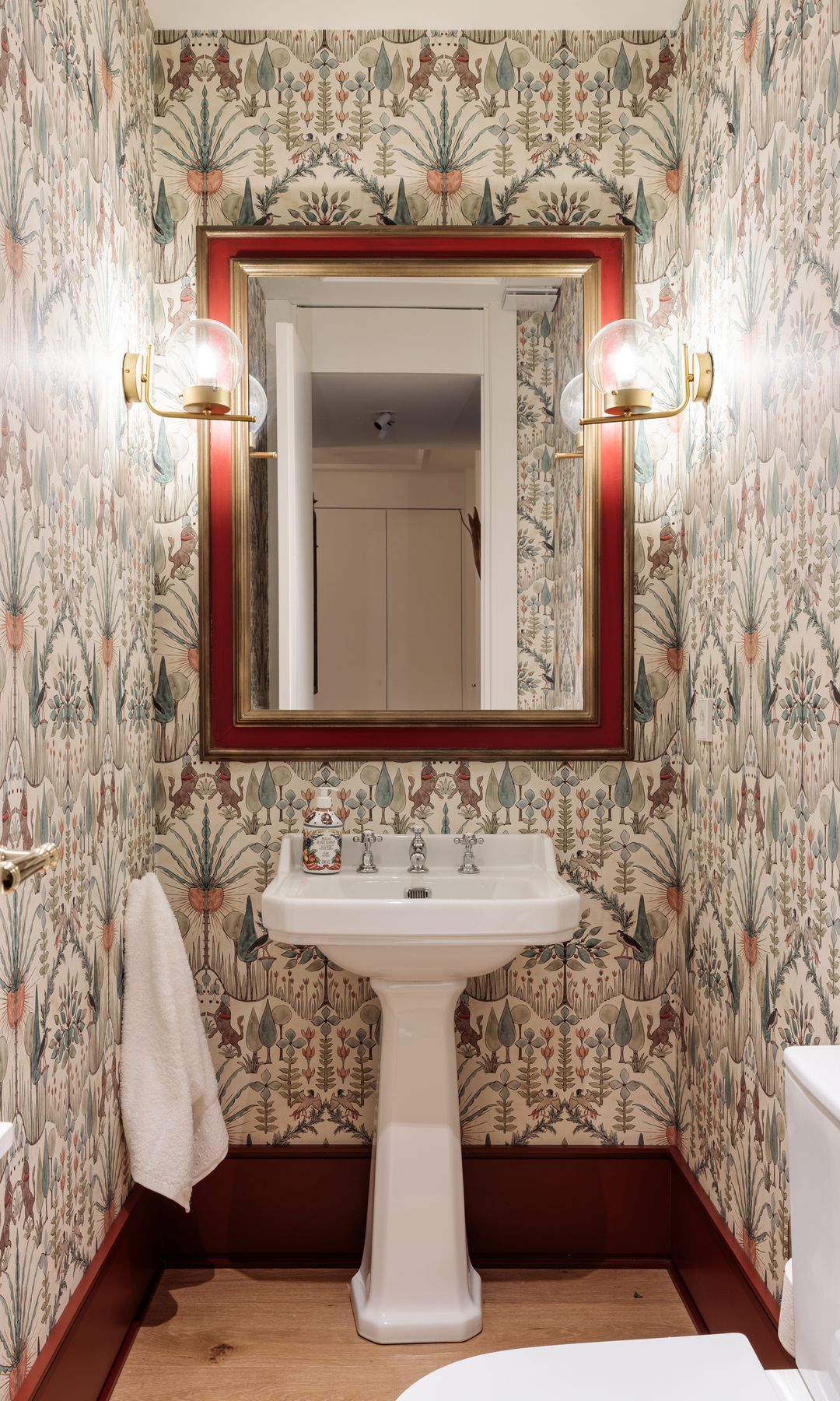 Baño con papel pintado, espejo con el marco de madera, apliques de pared encendidos, lavabo de pedestal