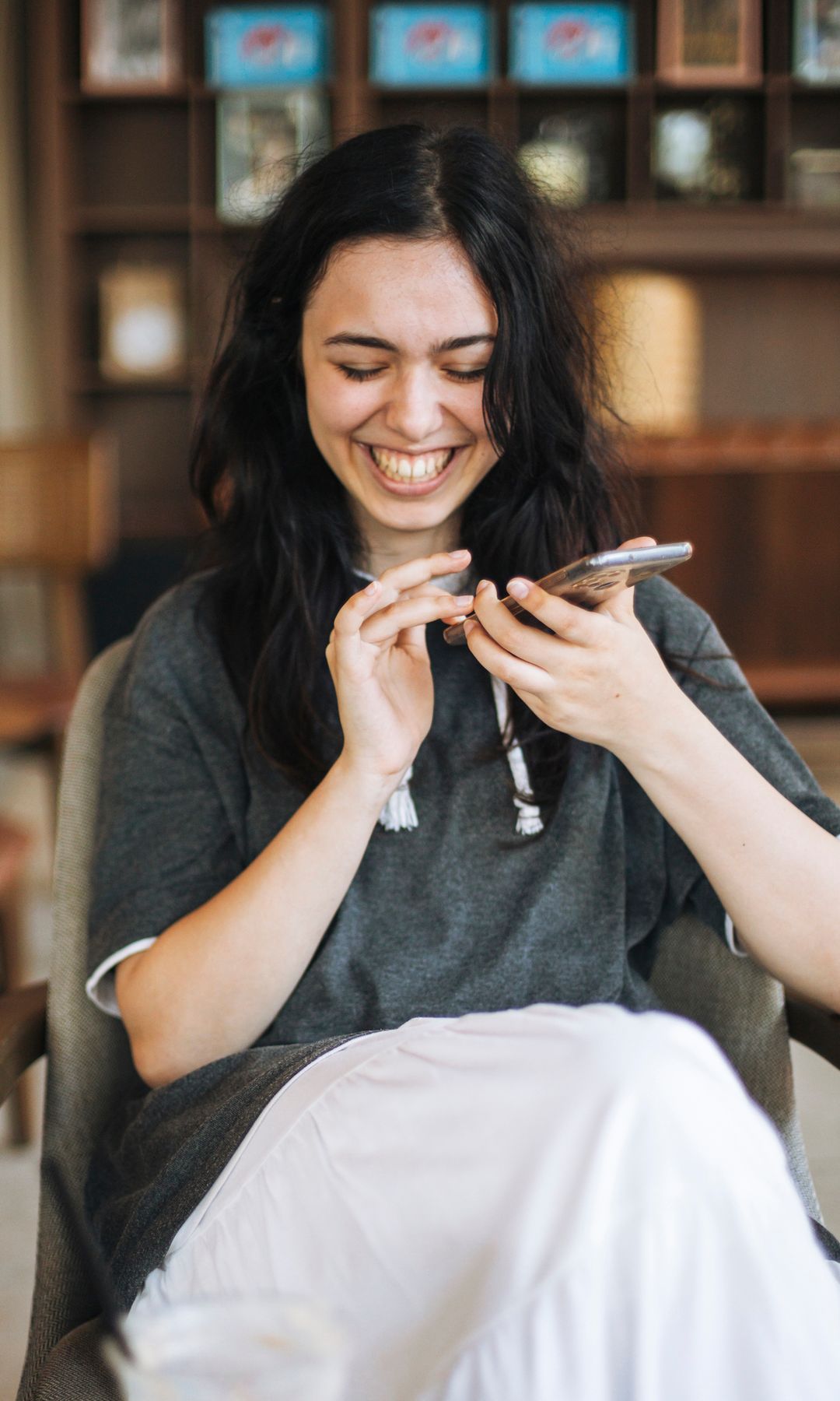 Mujer con cabello largo, riendo y usando un teléfono en una silla.