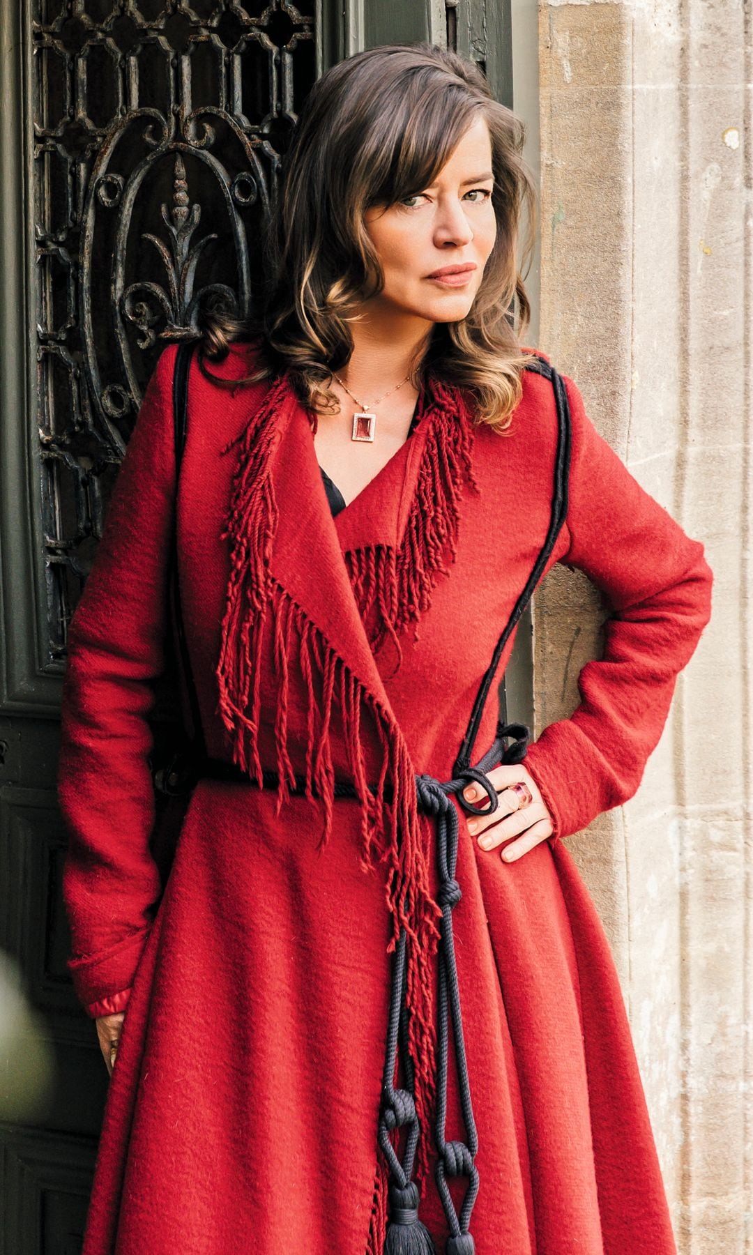 Jade Jagger posa junto a una puerta antigua, vestida con un abrigo rojo con flecos.