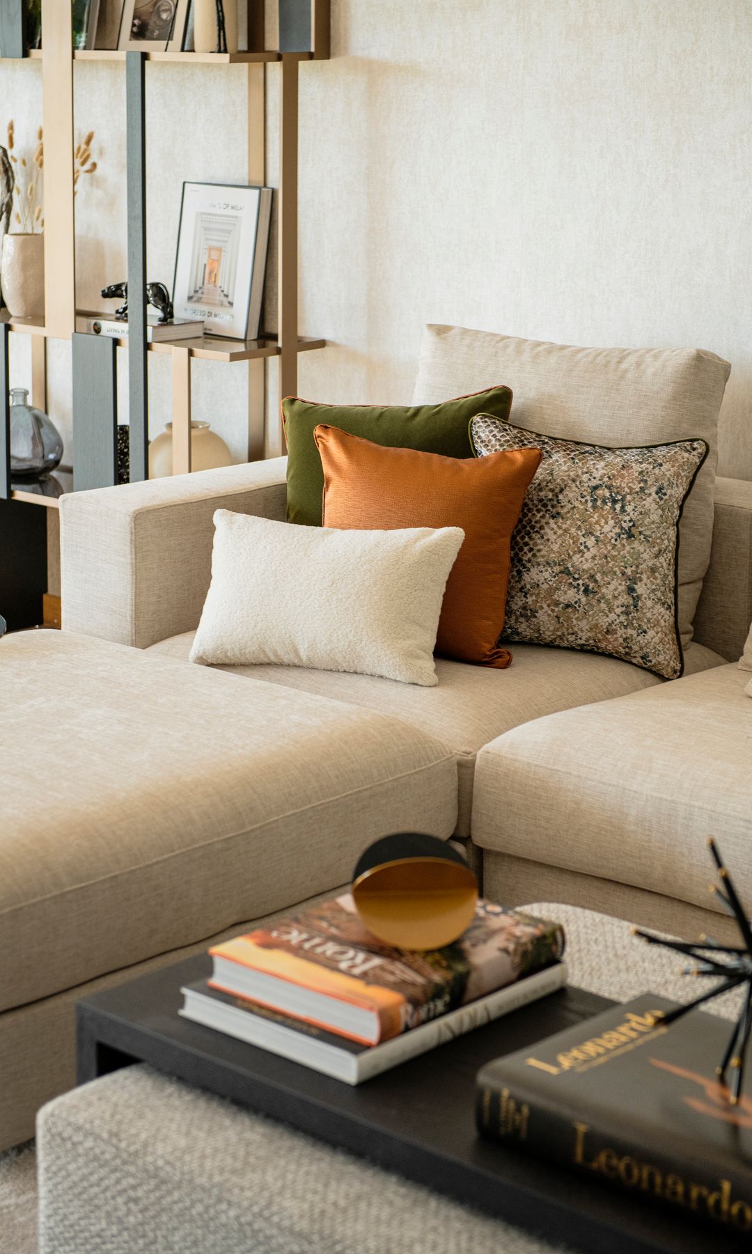 Salón con sofá beige, estantería con marcos y libros, mesa de centro tapizada con balda de madera negra con libros y adornos, cojines en el sofá