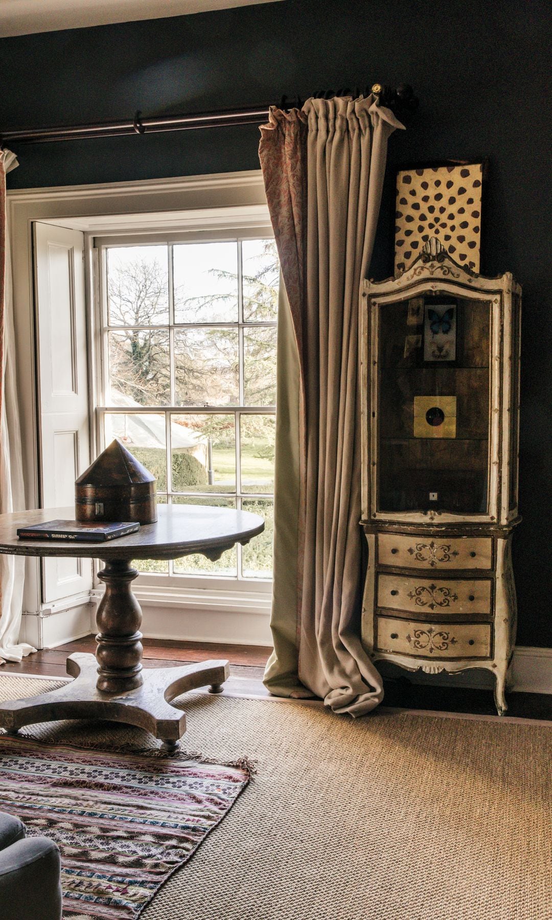 Habitación con ventana, cortinas, mesa redonda con libro y objeto decorativo, y mueble antiguo.