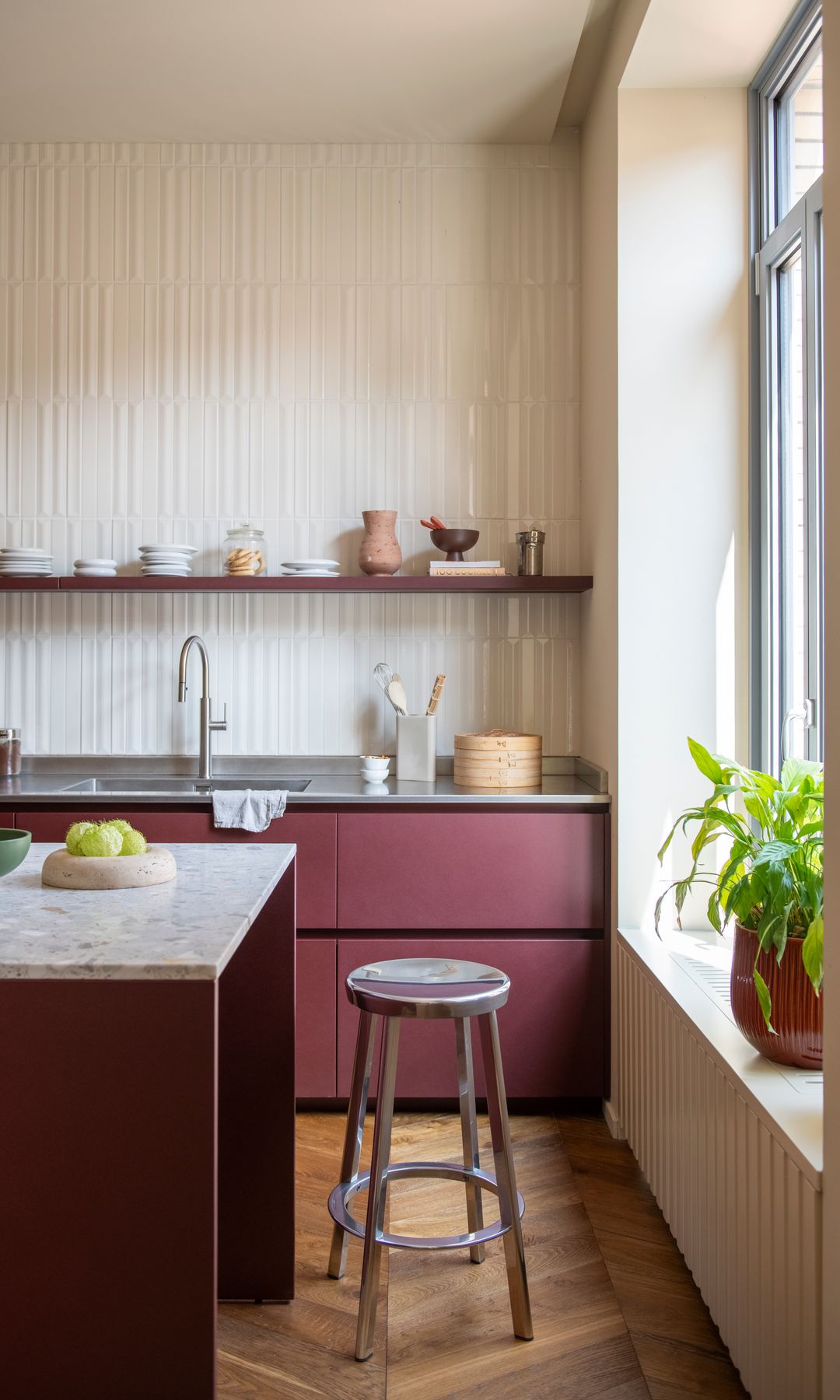 Cocina con isla, sin muebles superiores, muebles burdeos, ventanal y alféizar con plantas, paredes beiges, taburete metálico 