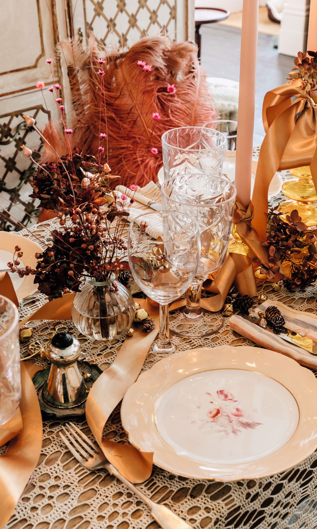 Mesa decorada con mantel de encaje, copas, cubiertos, y flores en jarrones.