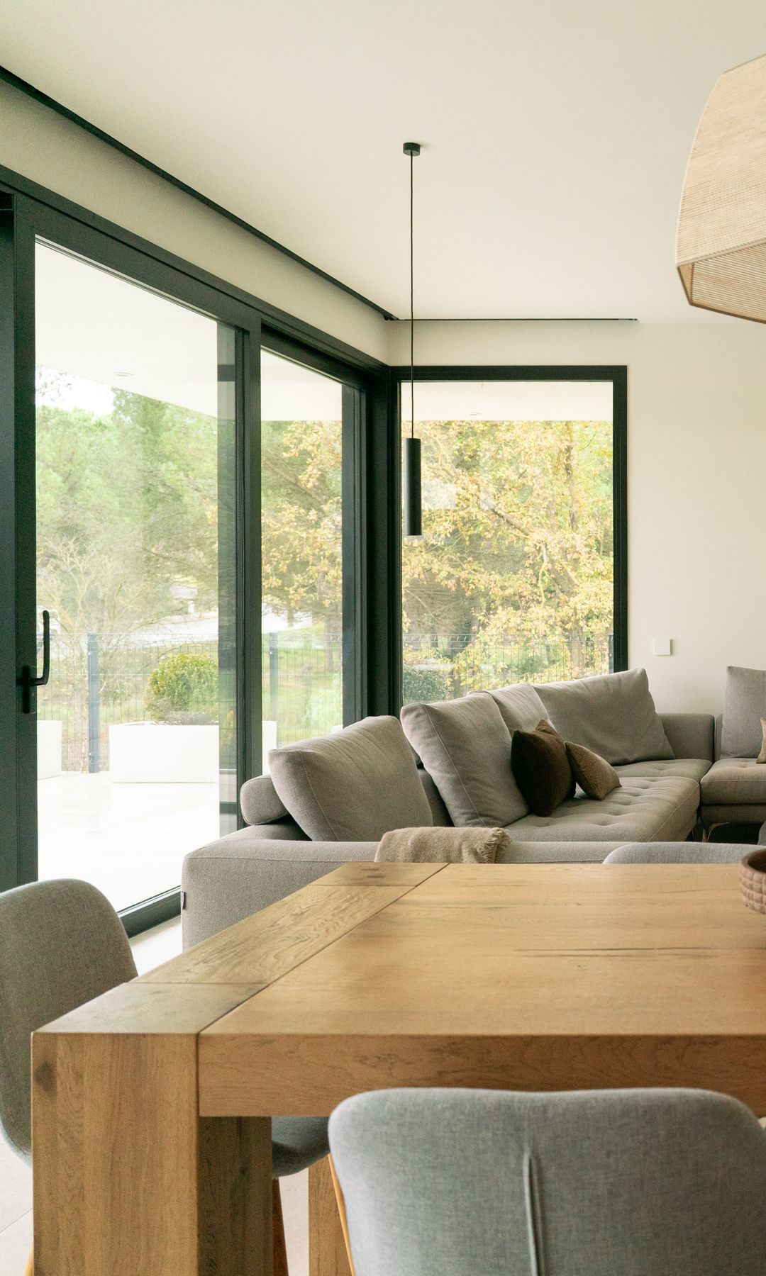 Salón comedor con chimenea moderna, ventanales con salida al jardín, mesa de madera, lámpara de techo de fibras vegetales, sillas tapizadas, sofá gris