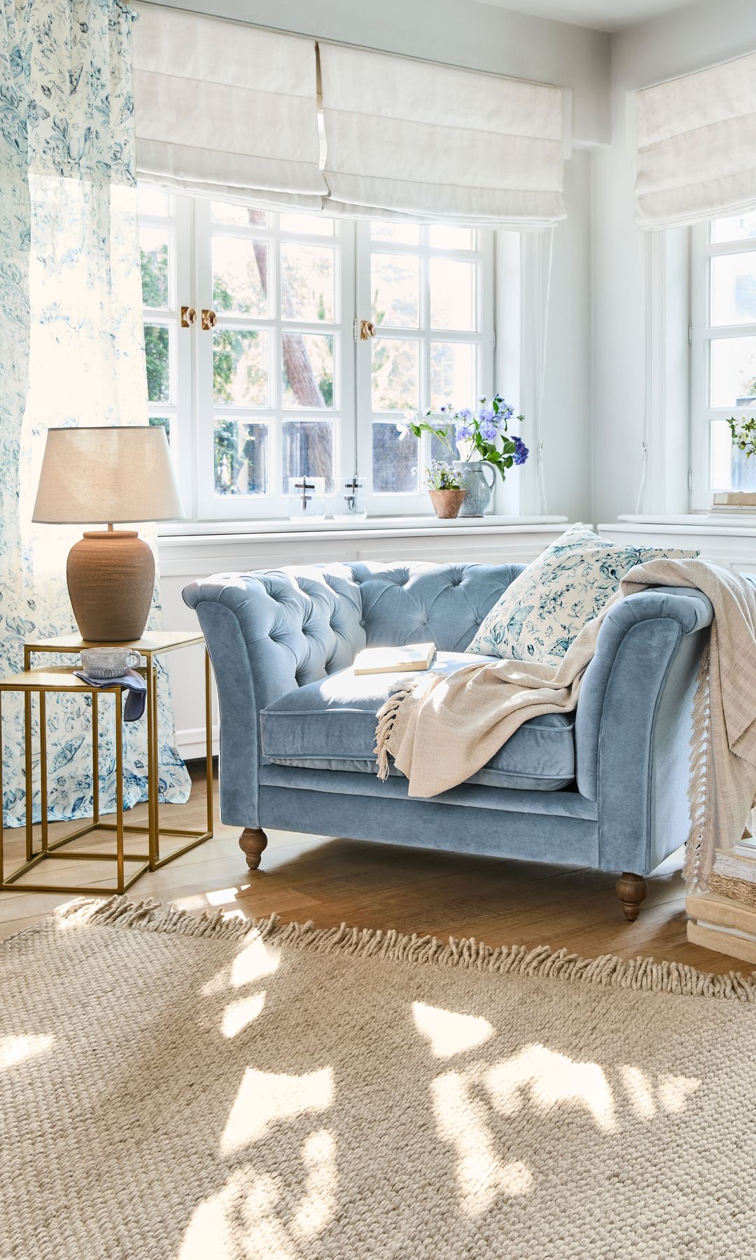 Rincón de lectura con sillón de terciopelo azul de capitoné, mesa auxiliar con lámpara de sobremesa, alfombra de fibras vegetales, estores blancos y cortinas de flores 