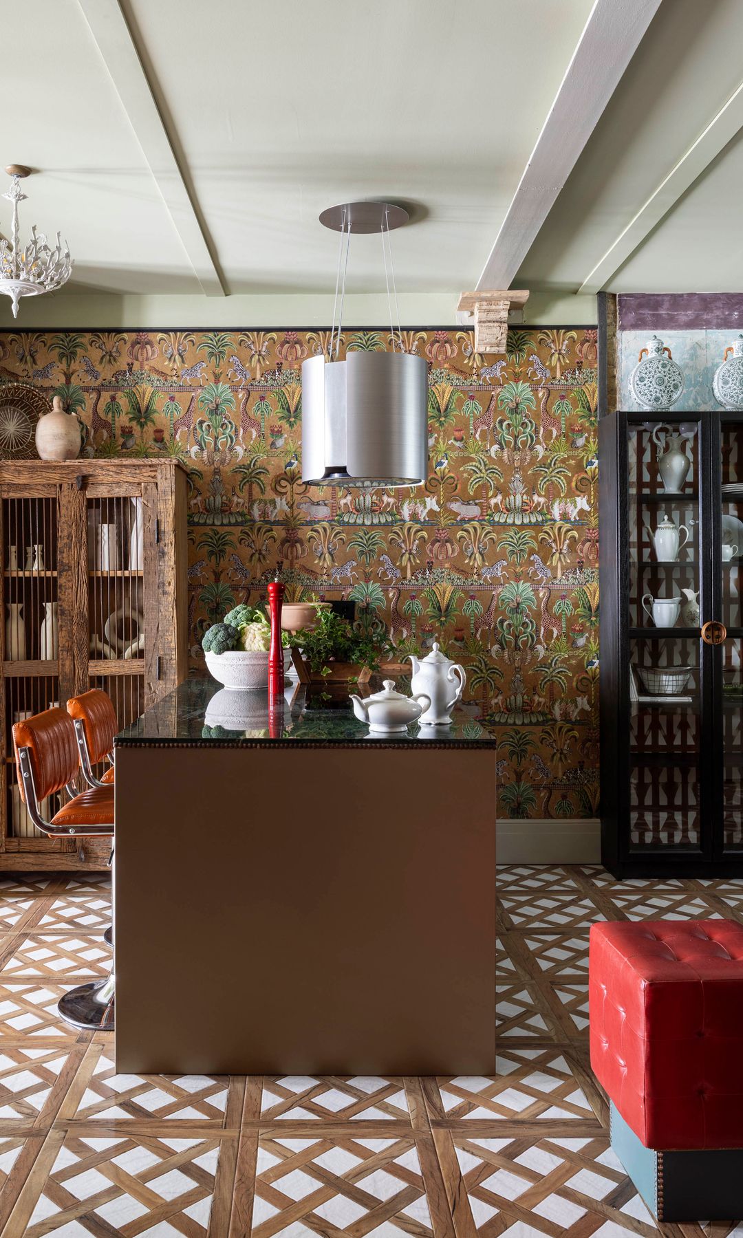 Isla de cocina con papel pintado, suelo de baldosas, taburetes de cuero, jarrones sobre la isla, vitrinas de madera y de color negro, campana de techo que es también lámpara 