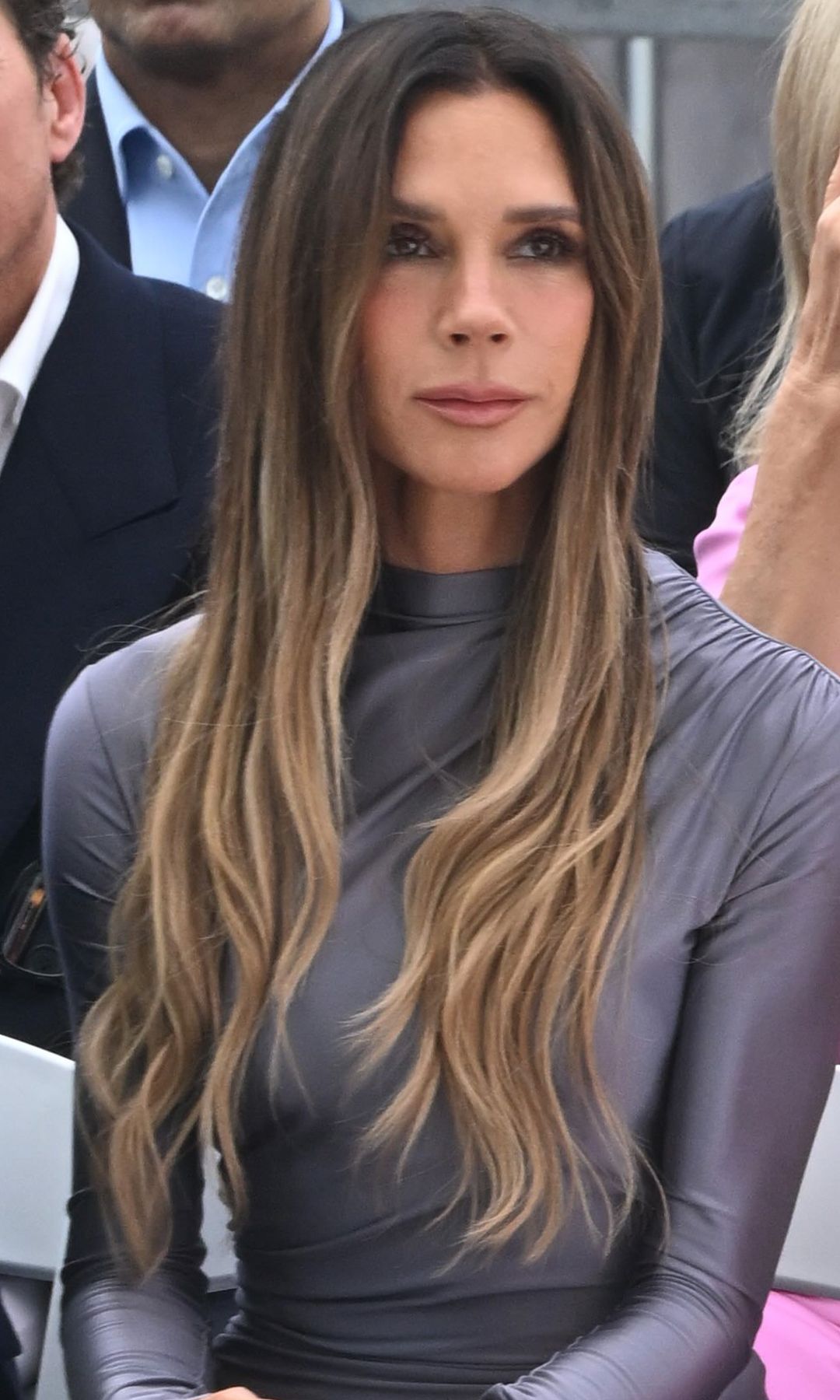 Victoria Beckham con vestido gris de cuello y mangas largas, pelo suelto y maquillaje natural