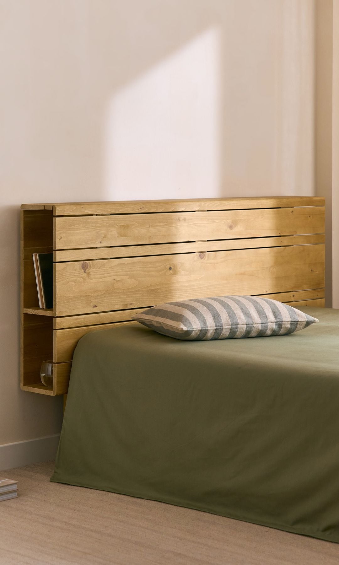 Dormitorio con ropa de cama verde, cabecero de madera con el lateral abierto y con libros, cojín y suelo de madera