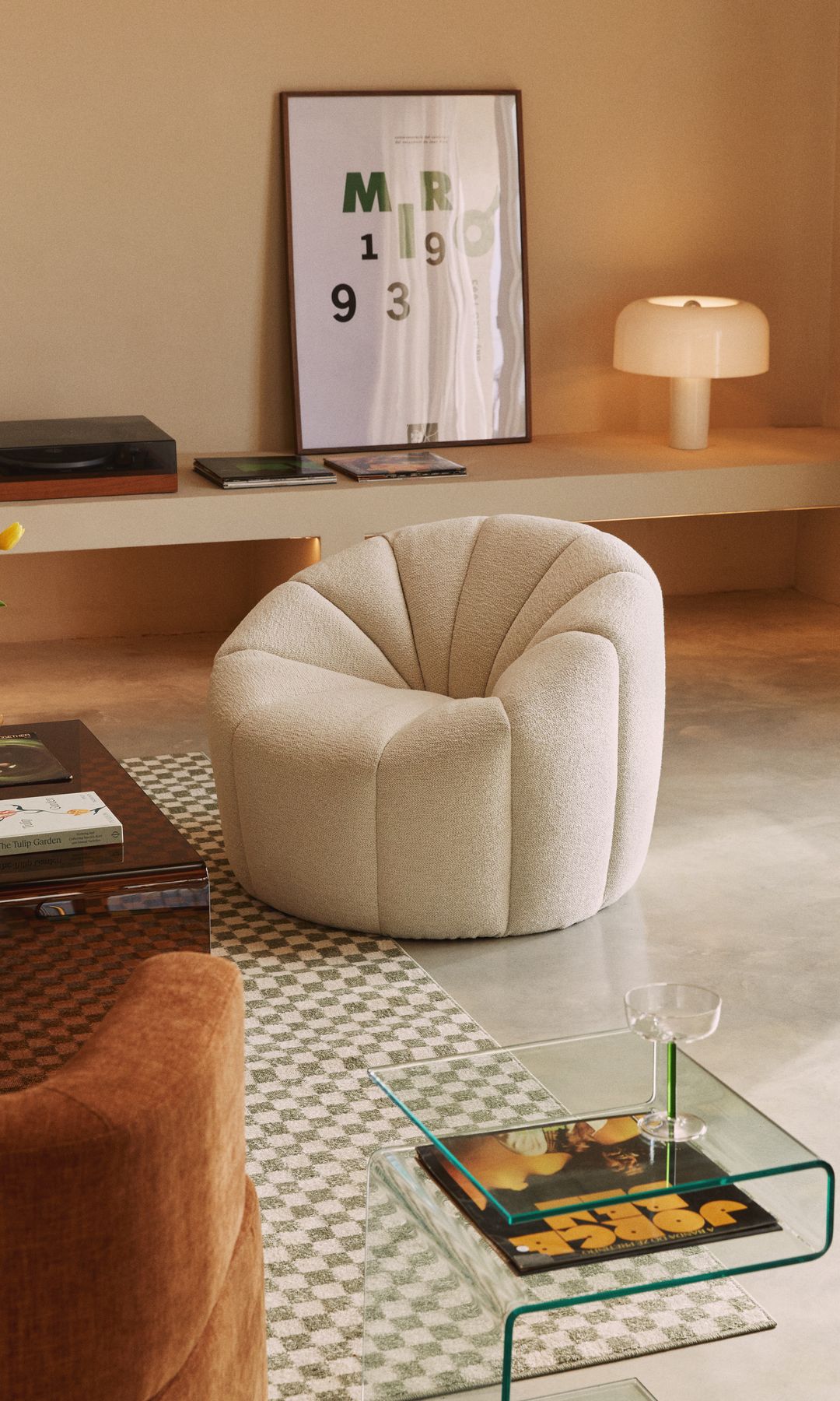 Salón de estilo nórdico con butaca curva blanca, mueble bajo con cuadro y lámpara de sobremesa, alfombra damero, sofá marrón