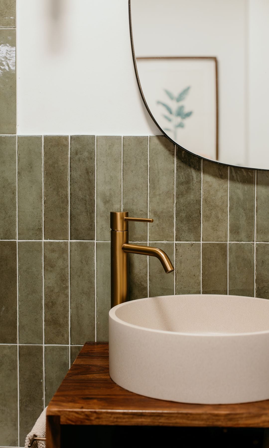 Baño con azulejos verdes, espejo, mueble de madera, grifería dorada, lavabo redondo sobre encimera