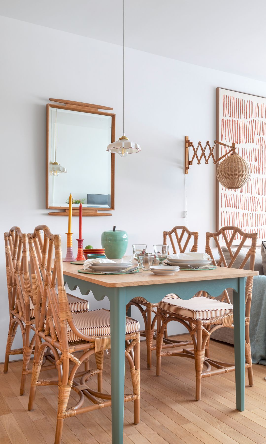 Comedor con mesa de madera con patas azules, sillas de comedor de mimbre, aplique de pared de fibras vegetales, suelo de madera, espejo con el marco de madera, candelabros de colores sobre la mesa
