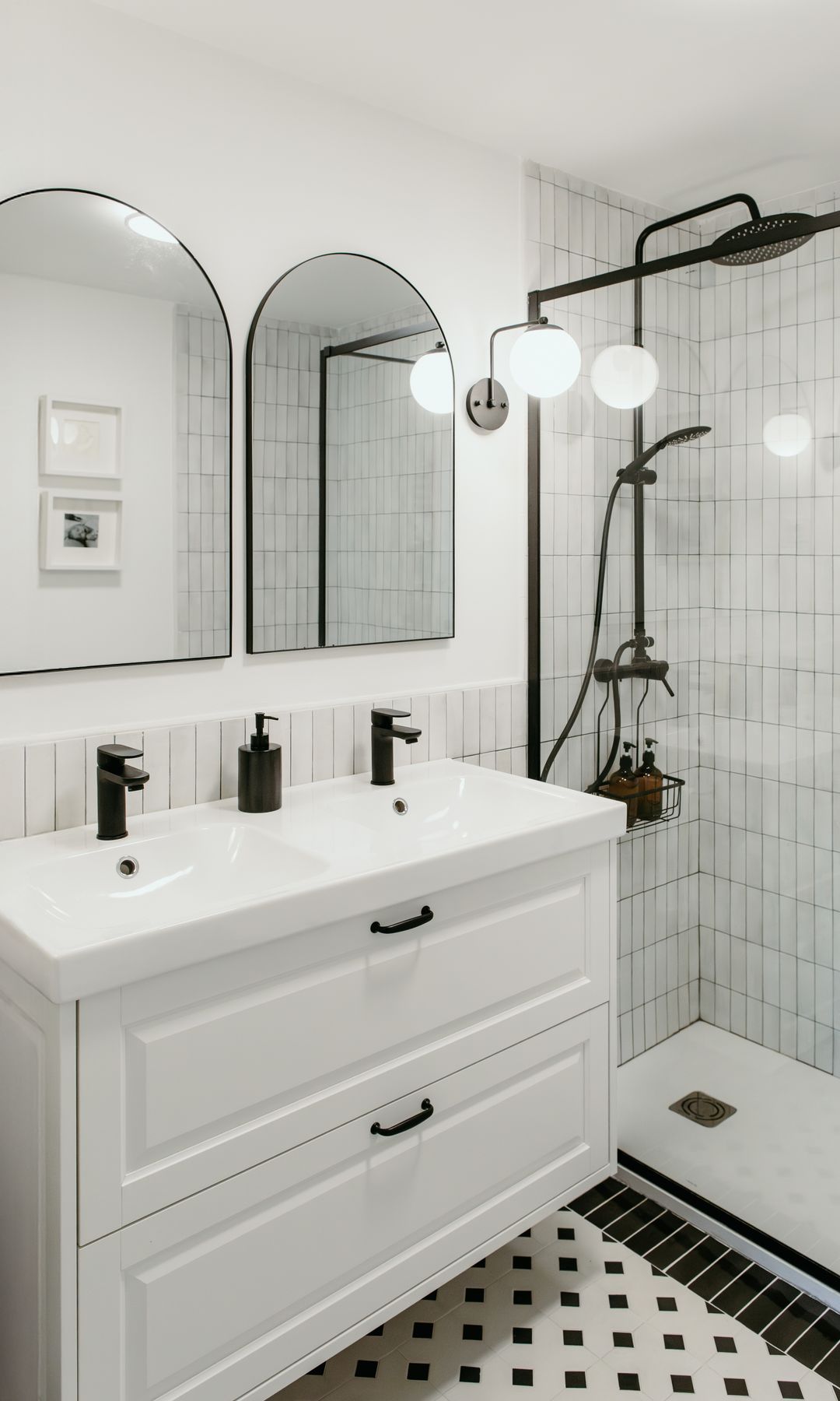 Baño con ducha en blanco y negro, mampara con perfiles negros, suelo blanco y negro, mueble bajolavabo suspendido blanco, dos espejos, grifería negra, aplique pared