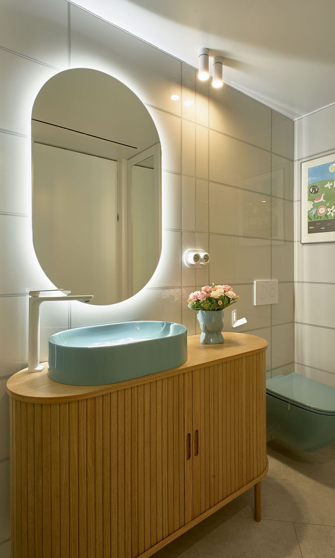 Baño con mueble de madera, espejo retroiluminado ovalado, inodoro y lavabo azul claro 