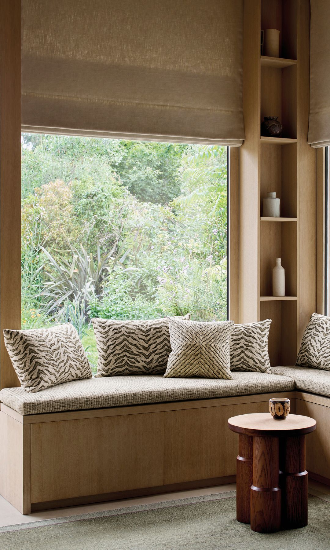 Banco de madera en L con capacidad de almacenaje, estores beige, cojines, estantería de madera, alfombra, mesa auxiliar