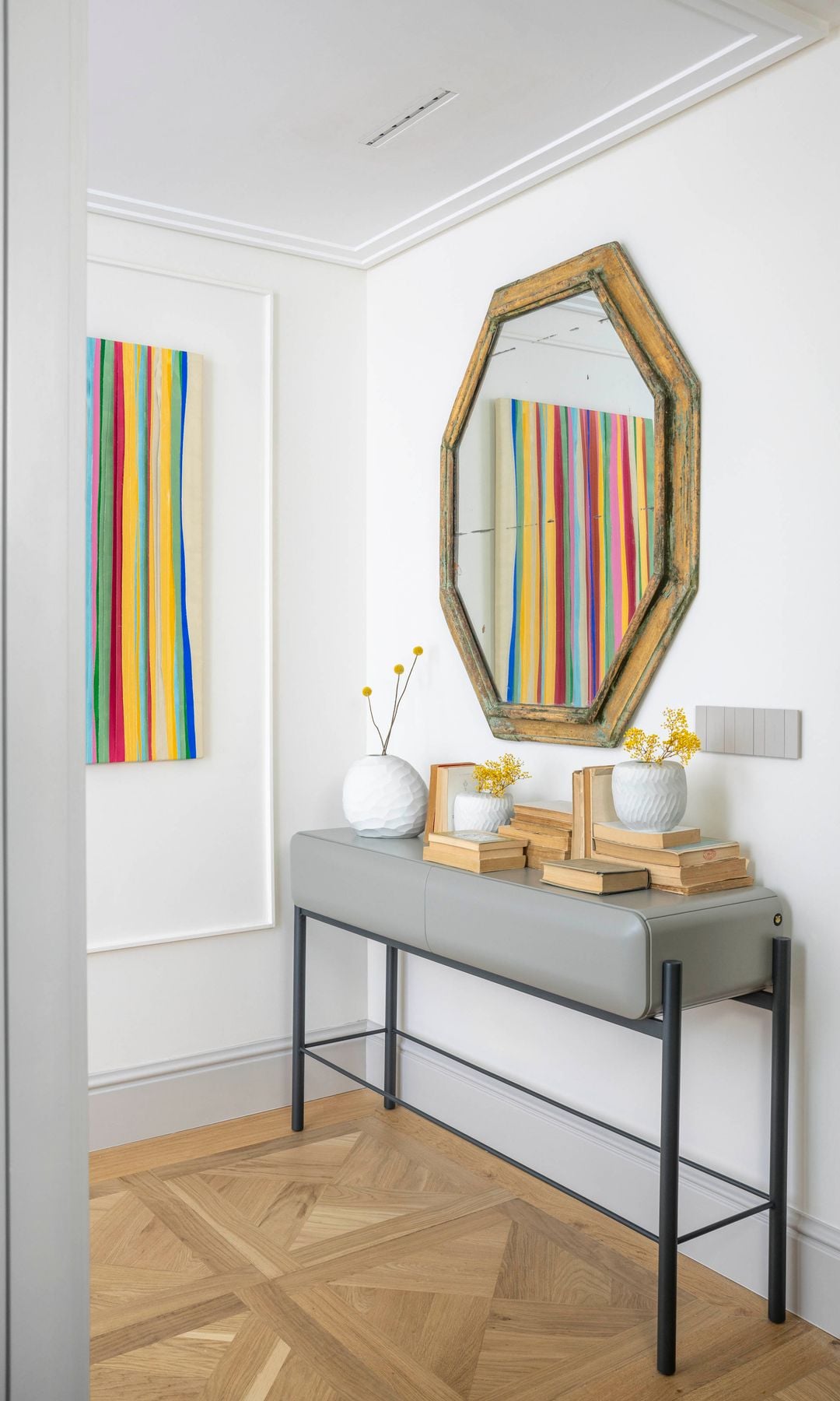 Recibidor con consola gris, espejo con el marco dorado, suelo de madera, libros y jarrones blancos con flores amarillas, cuadro de colores en la pared