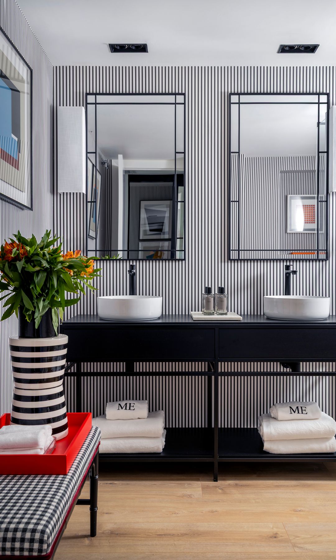 Baño en blanco y negro con papel pintado de rayas, dos espejos con el marco negro, banco de cuadros, bandeja roja, jarrón blanco y negro con flores 