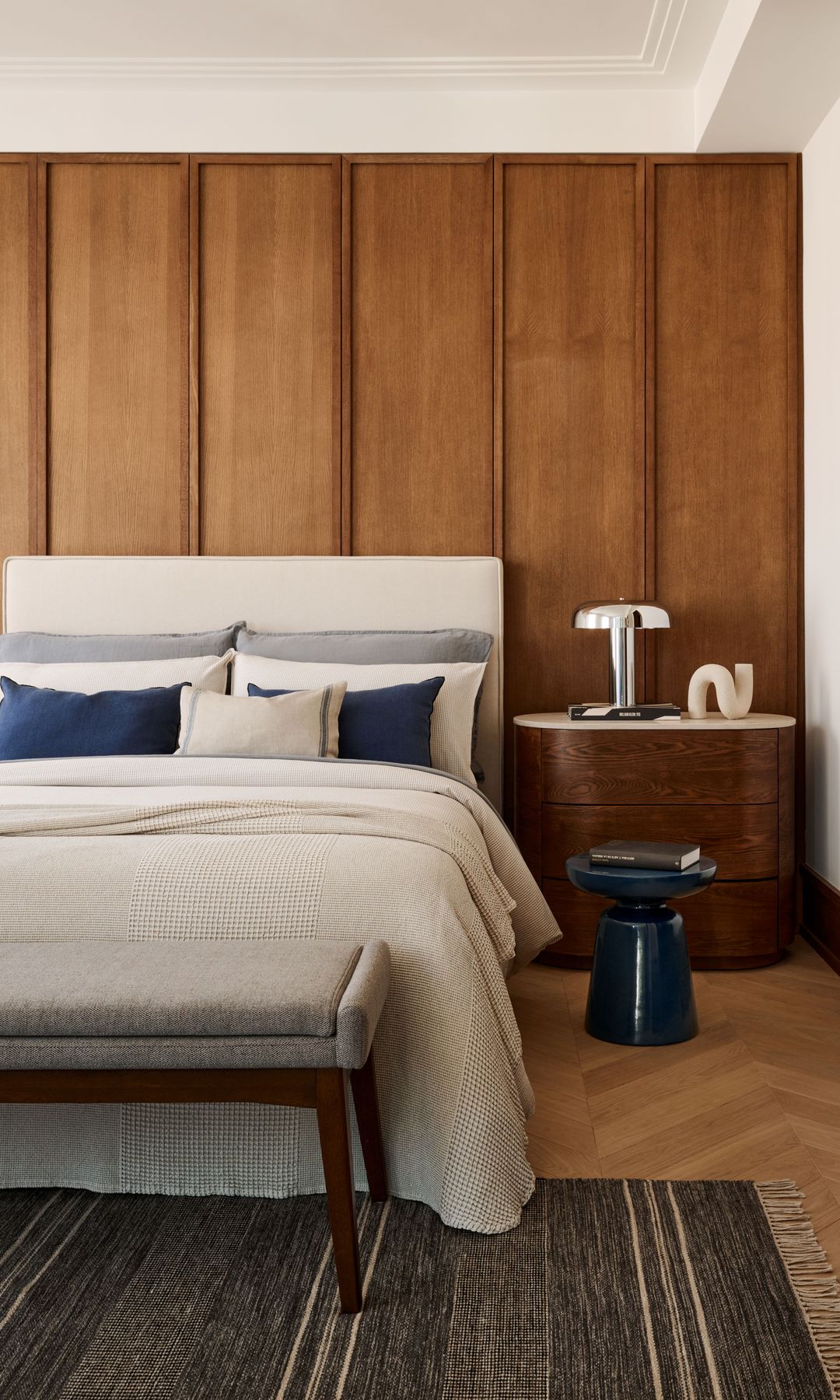 Dormitorio con panel de madera en la pared del cabecero, cabecero tapizado en color blanco, mesita de noche de madera con el sobre blanco, baldas de madera en hornacina, alfombra, ropa de cama beige