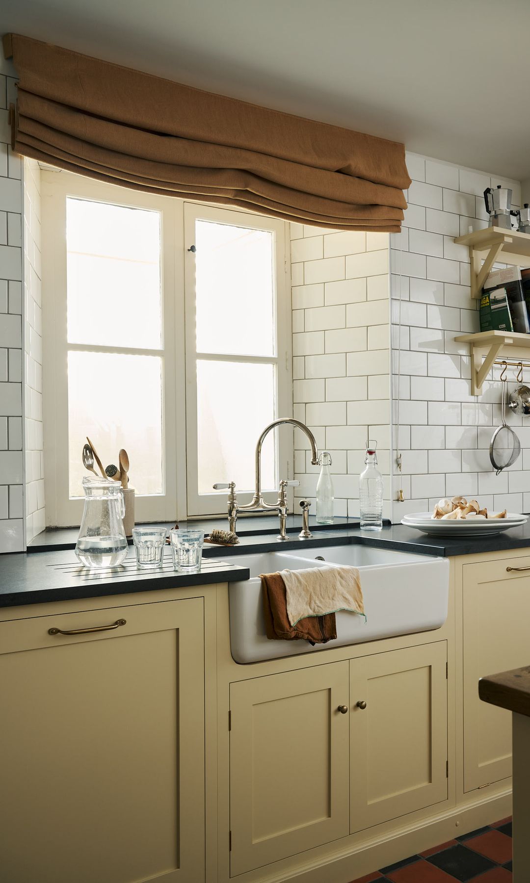 Cocina de estilo vintage con muebles de color vainilla, encimera negra, fregadero antiguo, grifo antiguo, ventana con perfiles de madera blancos, estor marrón 