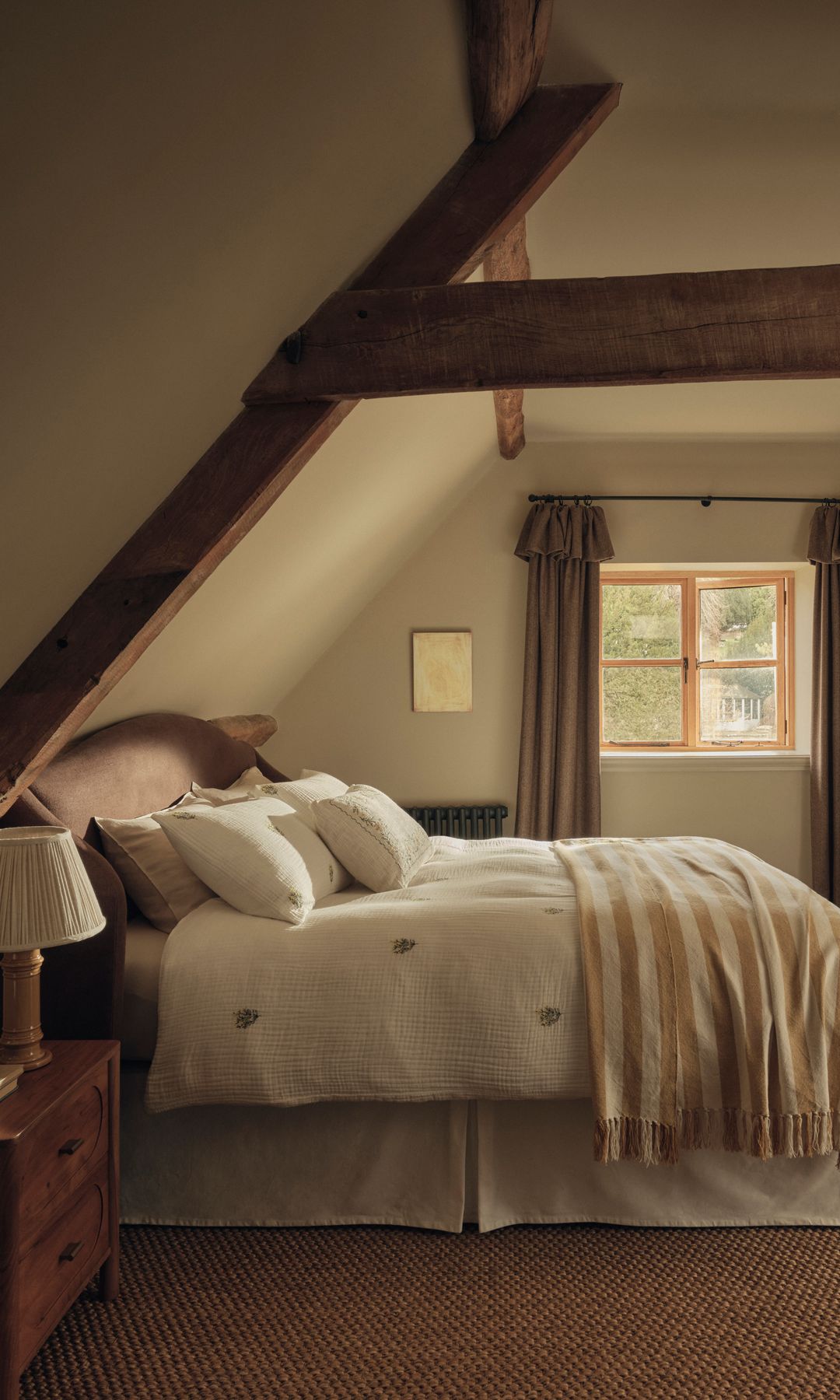 Dormitorio con el techo abuhardillado, viga de madera, ropa de cama blanca, mesita de noche de madera, lámpara de sobremesa 