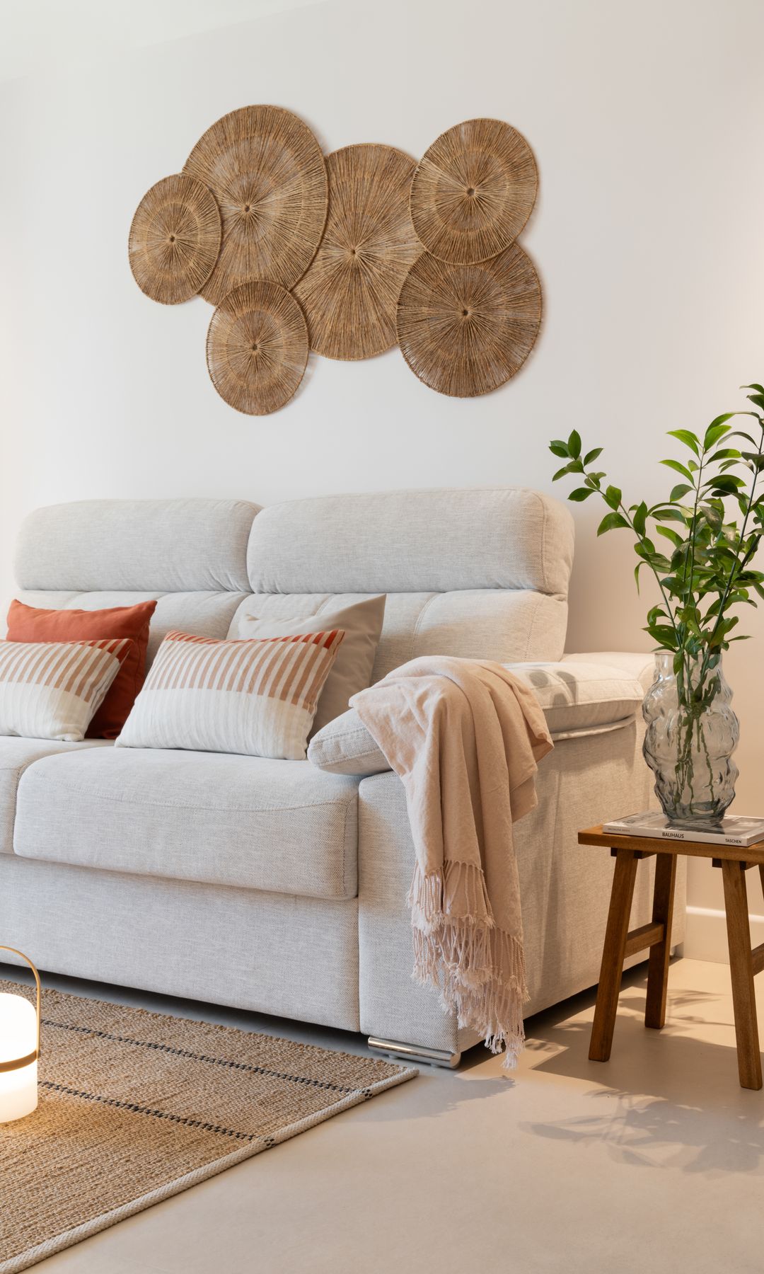 Salón con sofá blanco con pared del sofá con adornos de de fibras vegetales, alfombra de fritas naturales, taburete de madera, jarrón con planta
