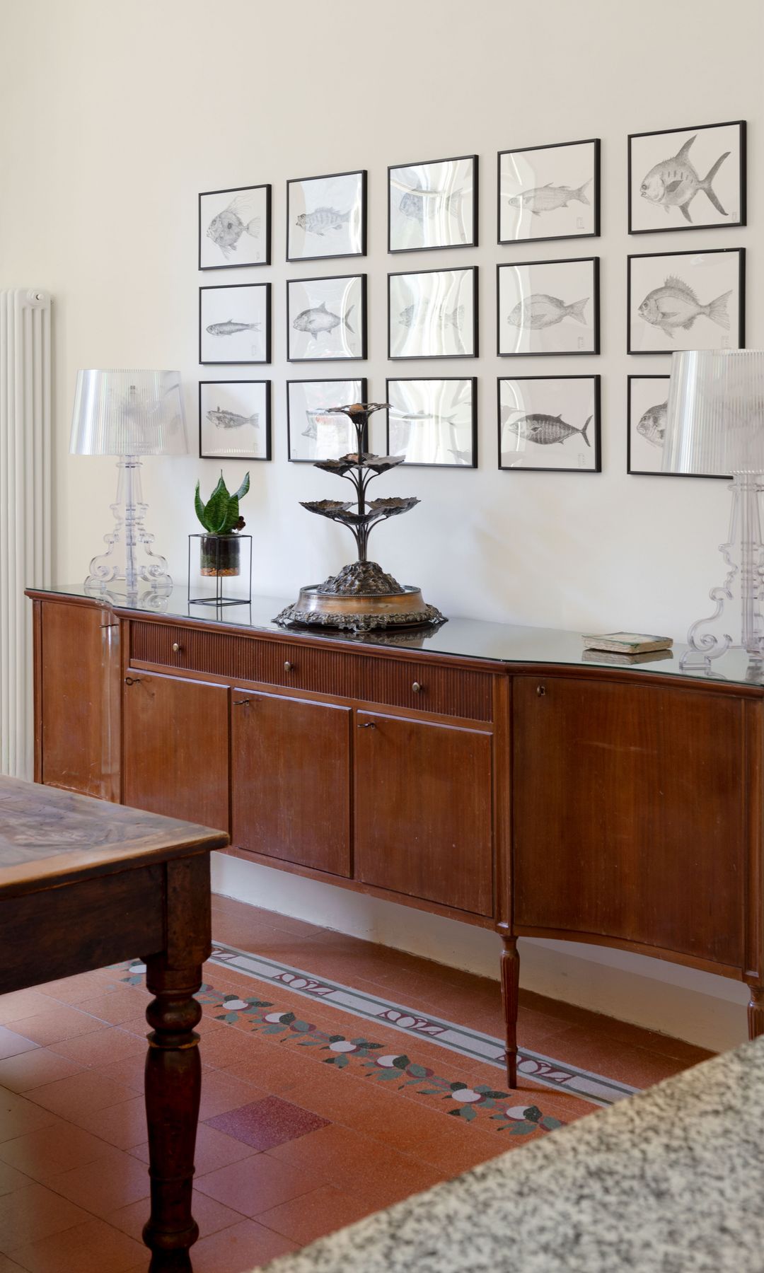 Aparador antiguo de madera oscura, con patas torneadas, lámparas de sobremesa de plástico transparente, cuadros en las paredes