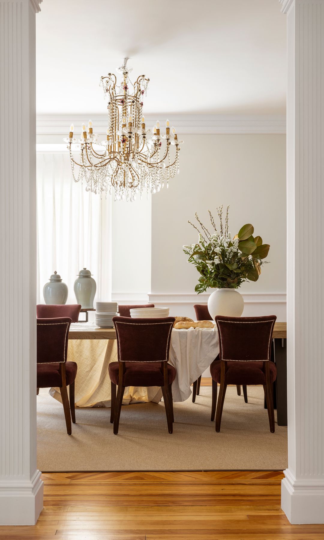 Comedor con lámpara de araña, sillas tapizadas, alfombra, mesa con mantel, jarrones con flores verdes, vajilla blanca sin colocar