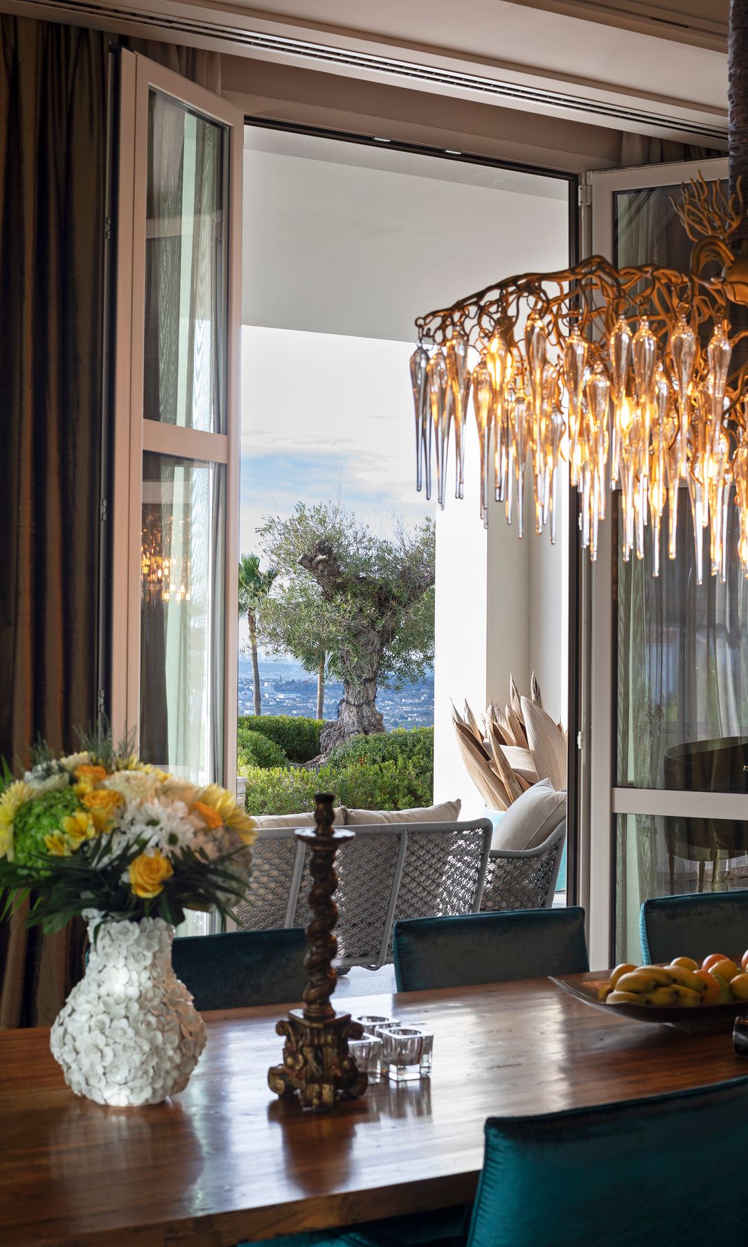 Comedor con ventanal con salida al porche, mesa de madera, lámpara de techo, sillas tapizadas, jarrón de cerámica blanca con flores, vela, frutero